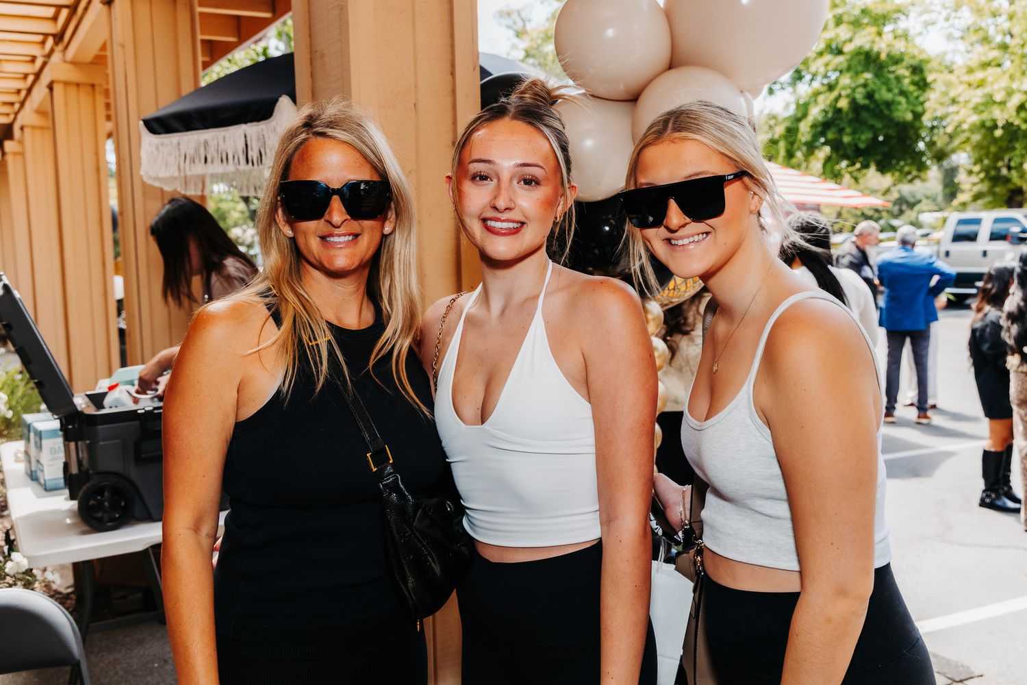 Three young blonde women wearing sunglasses pose together outdoors near balloons at a sunny outdoor event.