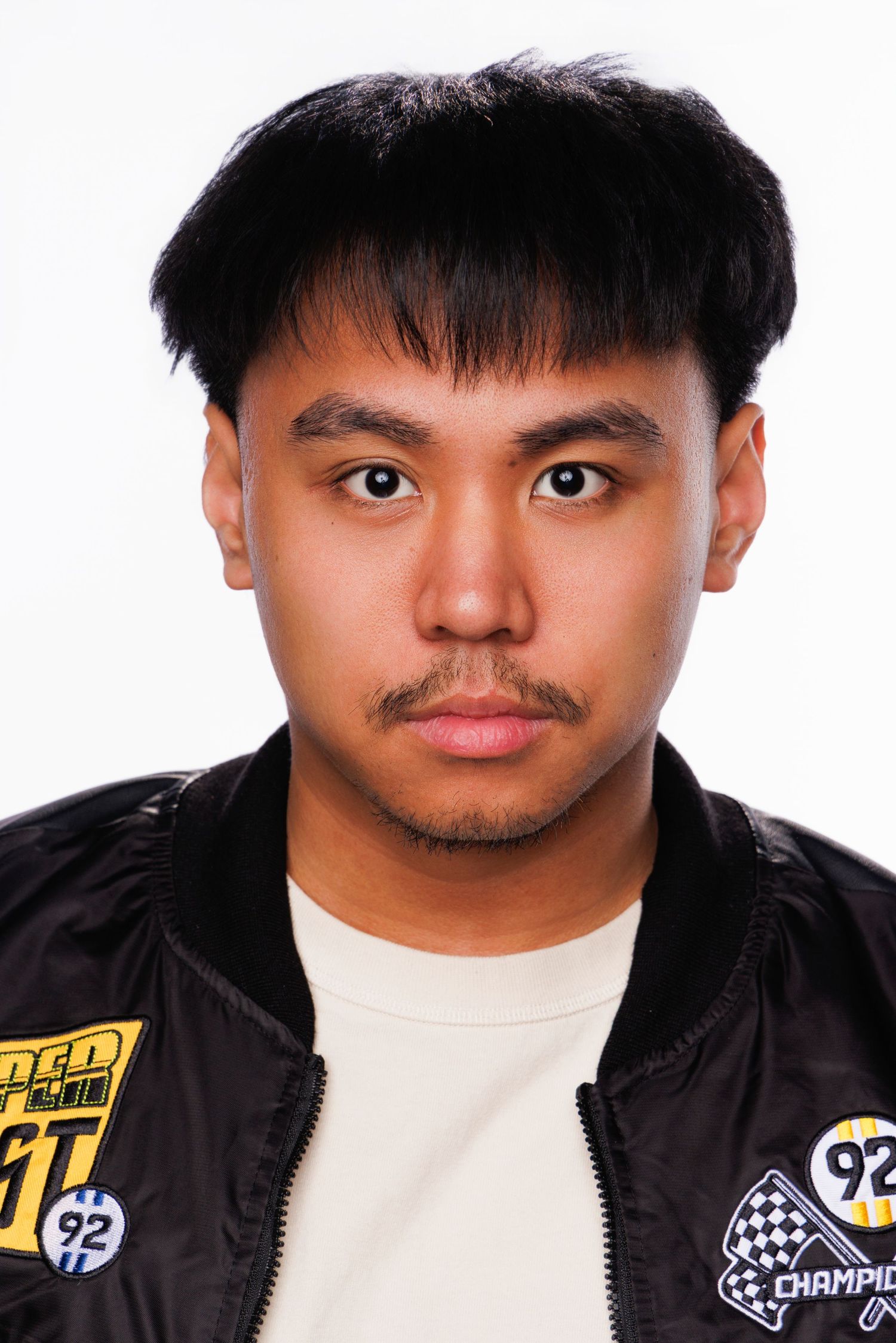 Young Asian man with black hair wearing a racing-style bomber jacket, posing against a white background.