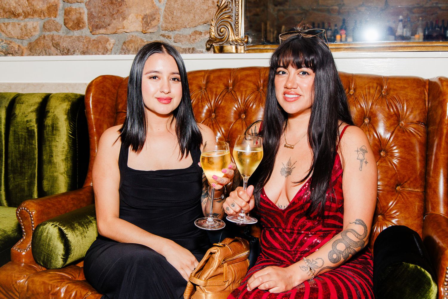 Two women sitting on a tufted leather sofa holding white wine glasses at a social event.
