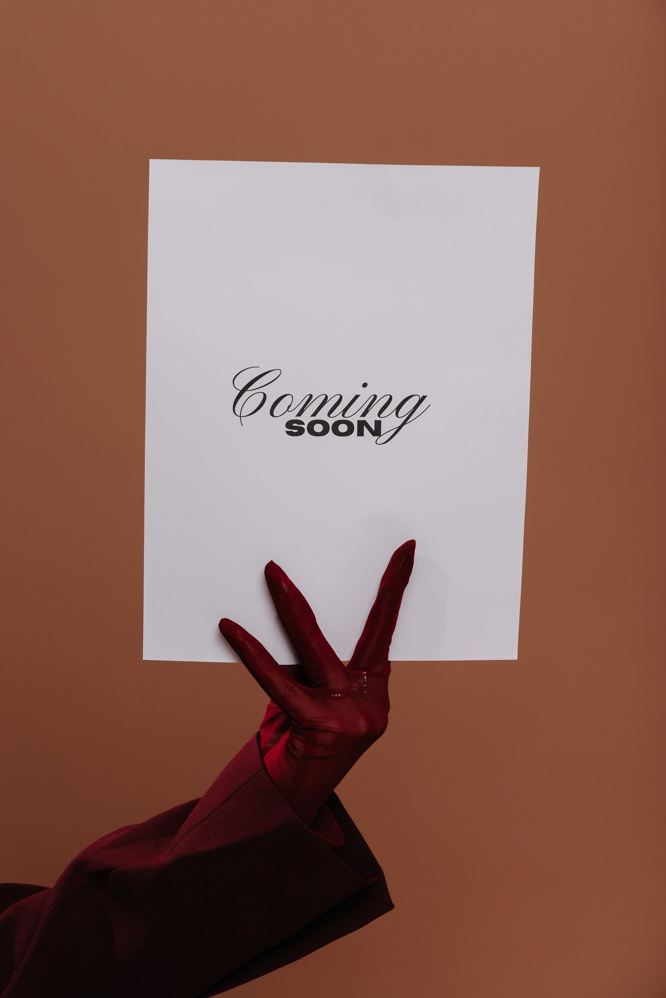 A red-gloved hand holds a white card displaying Coming Soon text against a brown background.