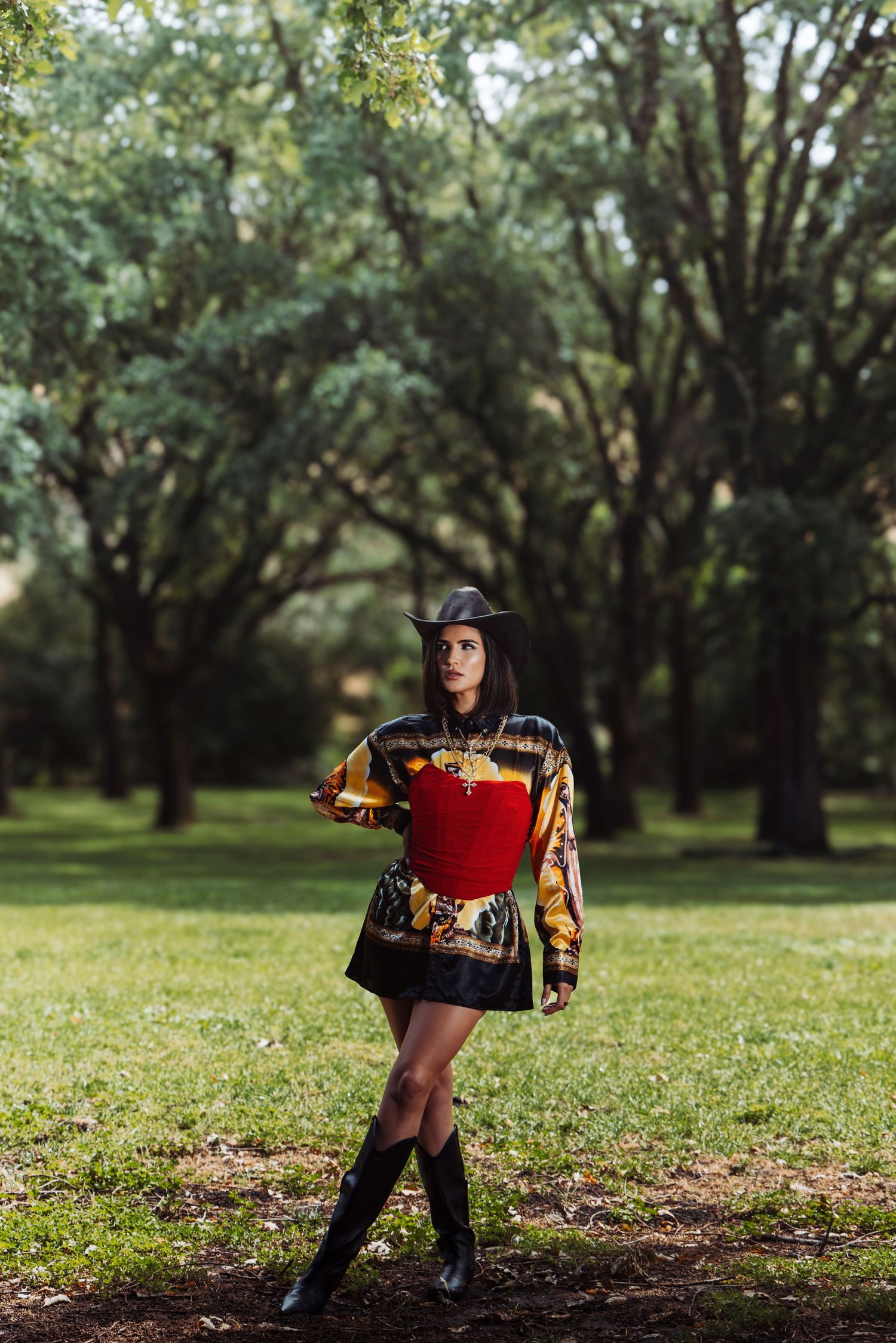 Woman in red corset, printed jacket, mini skirt, cowboy hat, and boots posing in a lush green park.