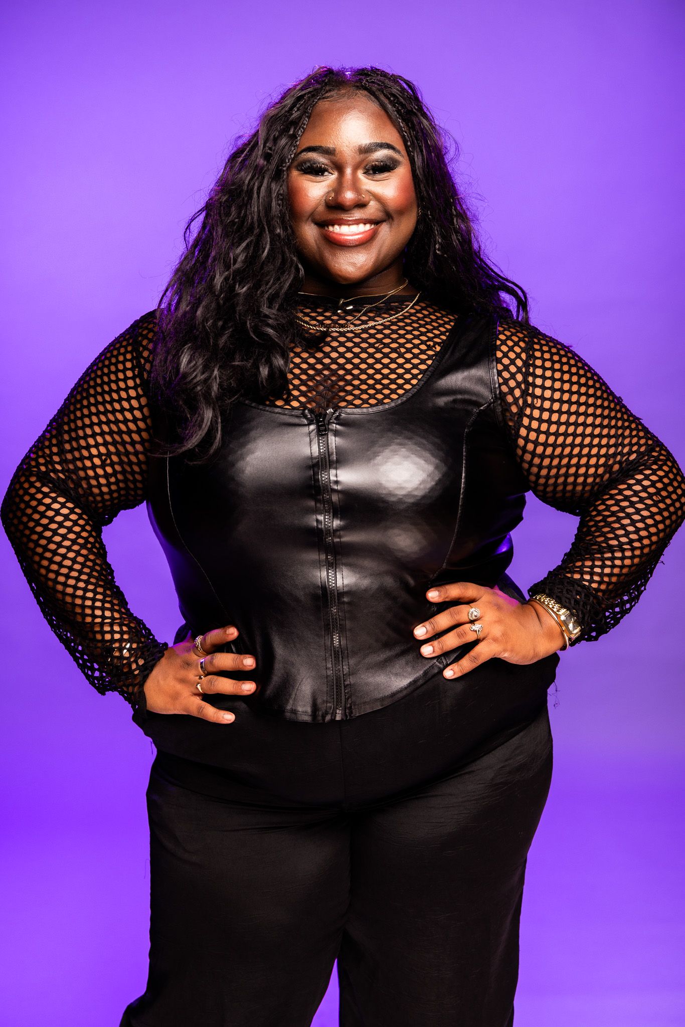 Smiling Black woman in black leather corset and fishnet top posing with hands on hips against purple background.
