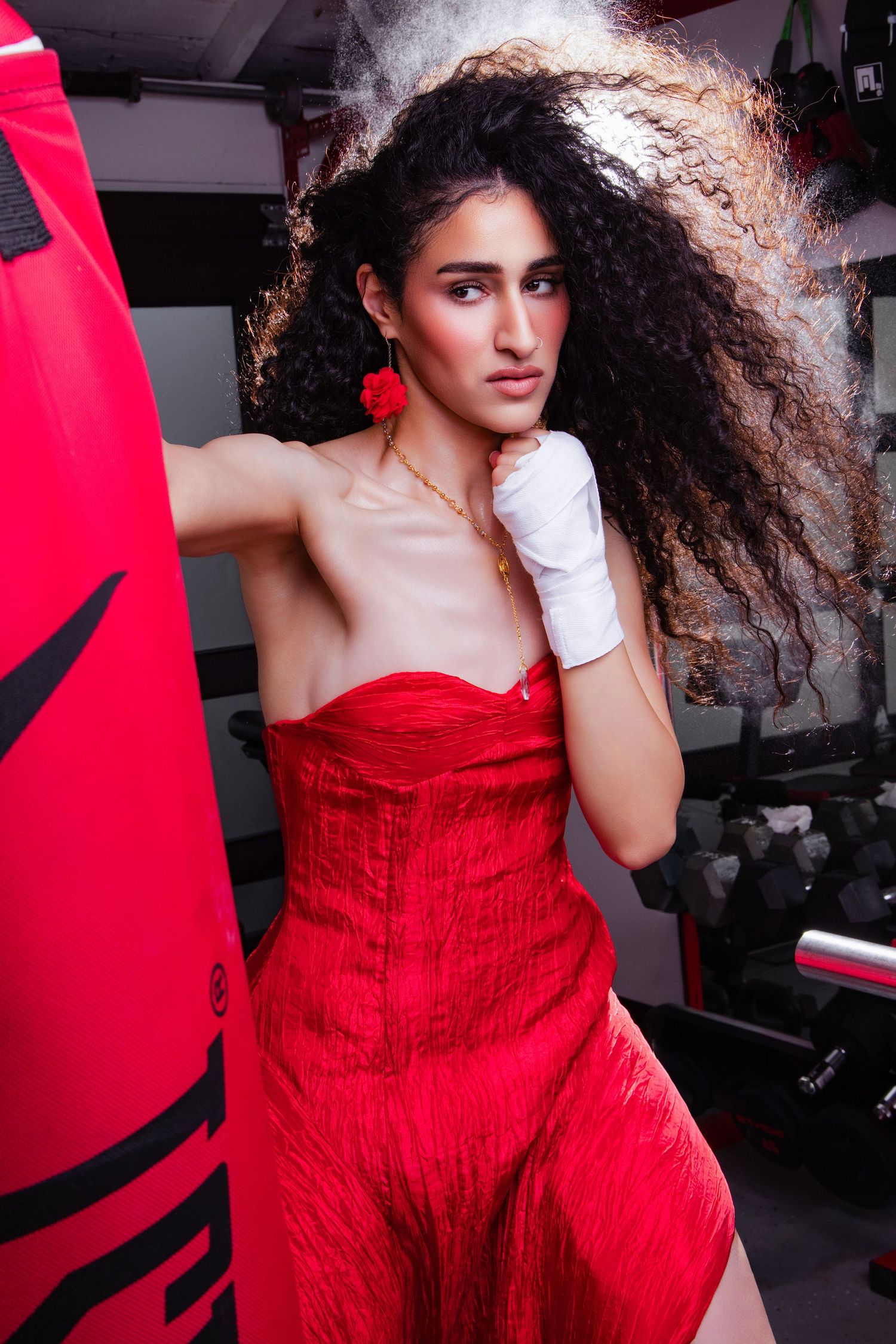 Woman in red strapless dress and white boxing gloves poses beside a red punching bag in a gym.