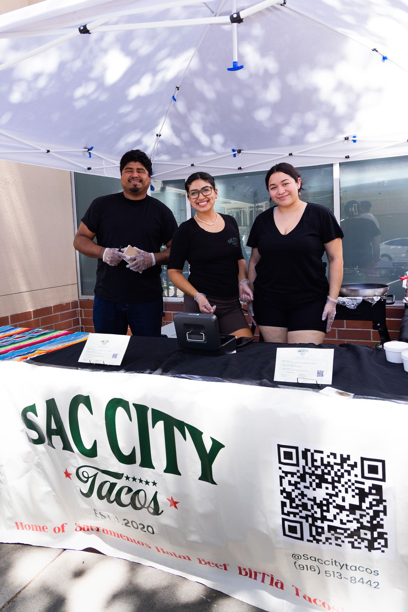 Three smiling staff members in black shirts stand behind a Sac City Tacos booth at an outdoor food event.