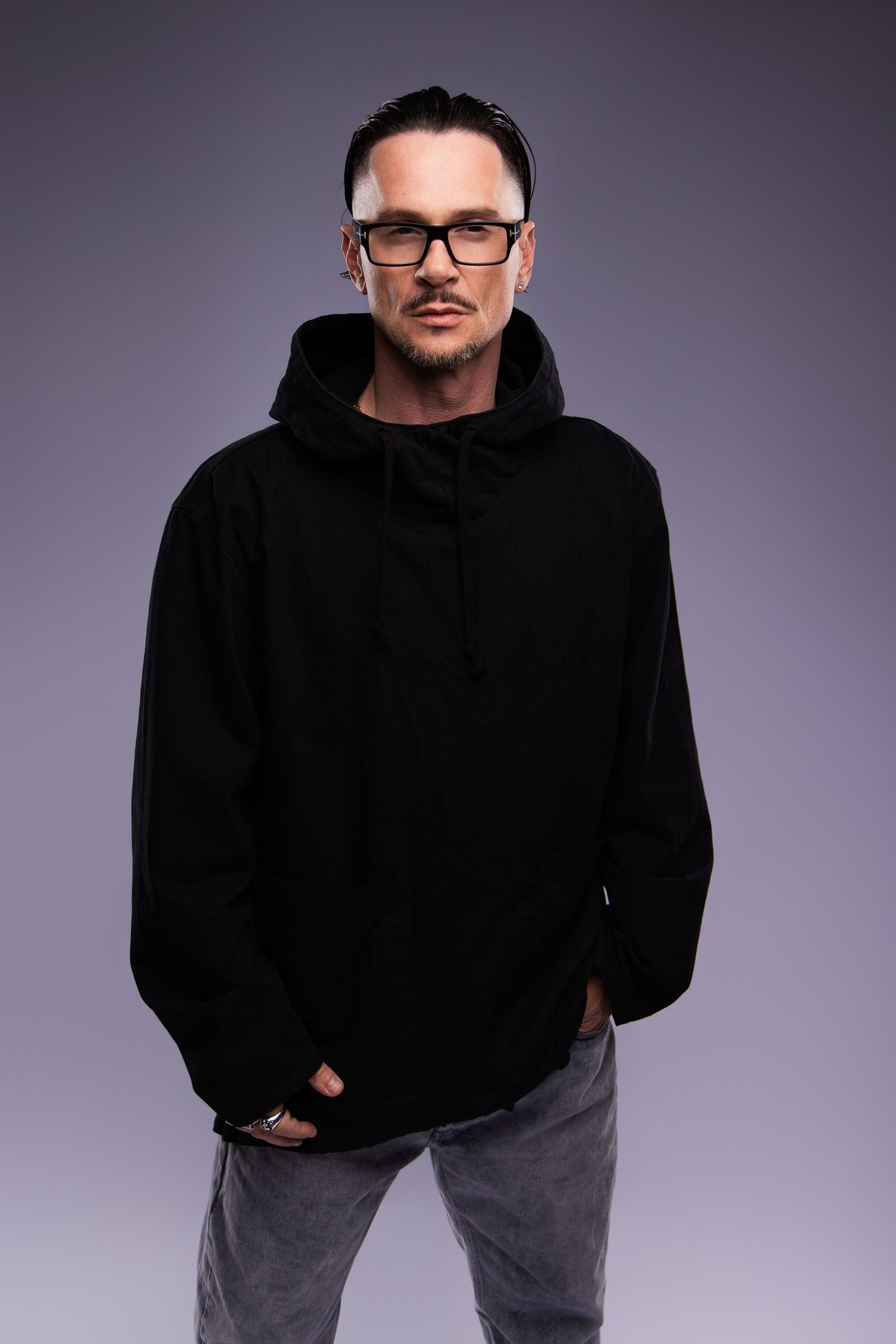 Man in black hoodie and glasses posing confidently against gray background.