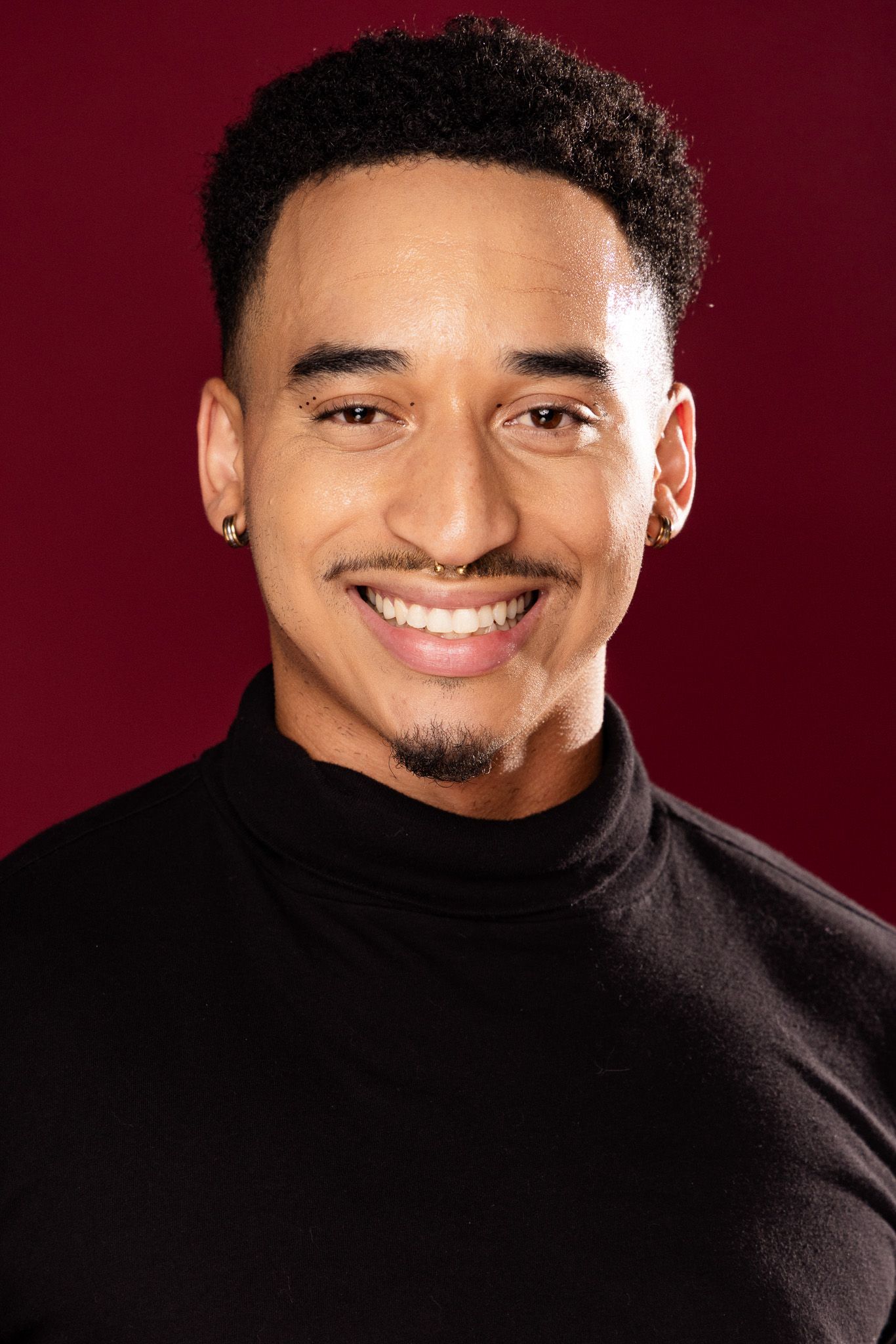 A smiling performer wearing a black turtleneck sweater poses against a burgundy background in a series of headshots.