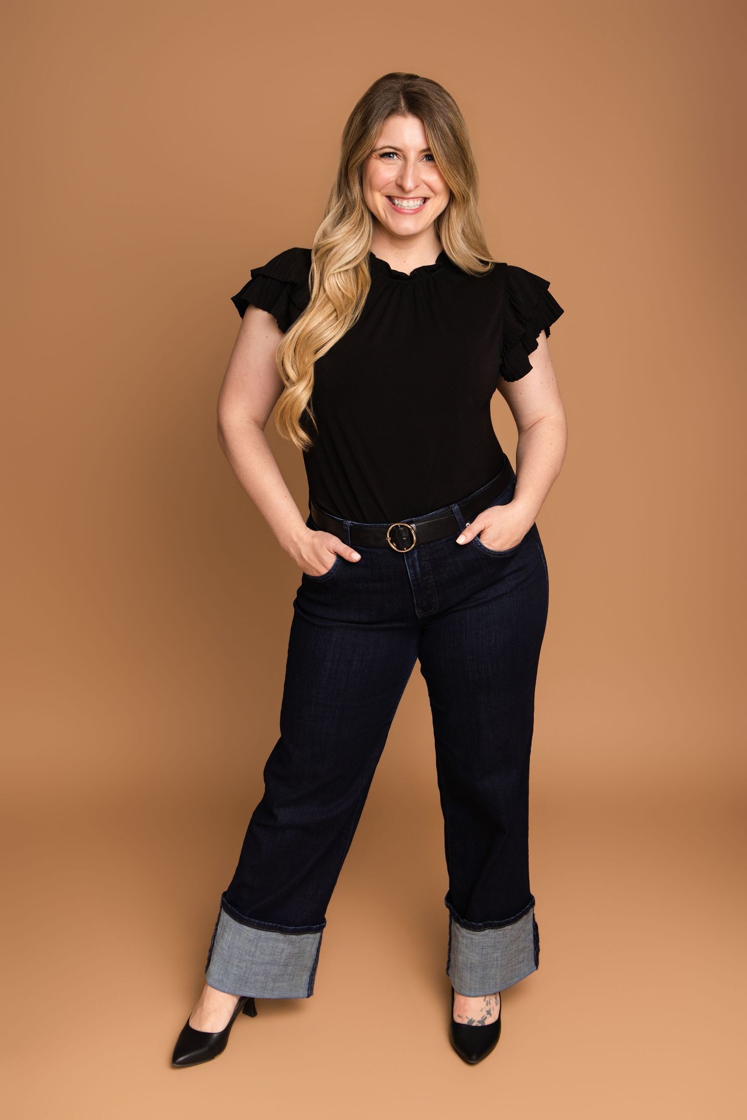 A person in a black ruffled sleeve top and wide-leg cuffed jeans poses against a brown backdrop.