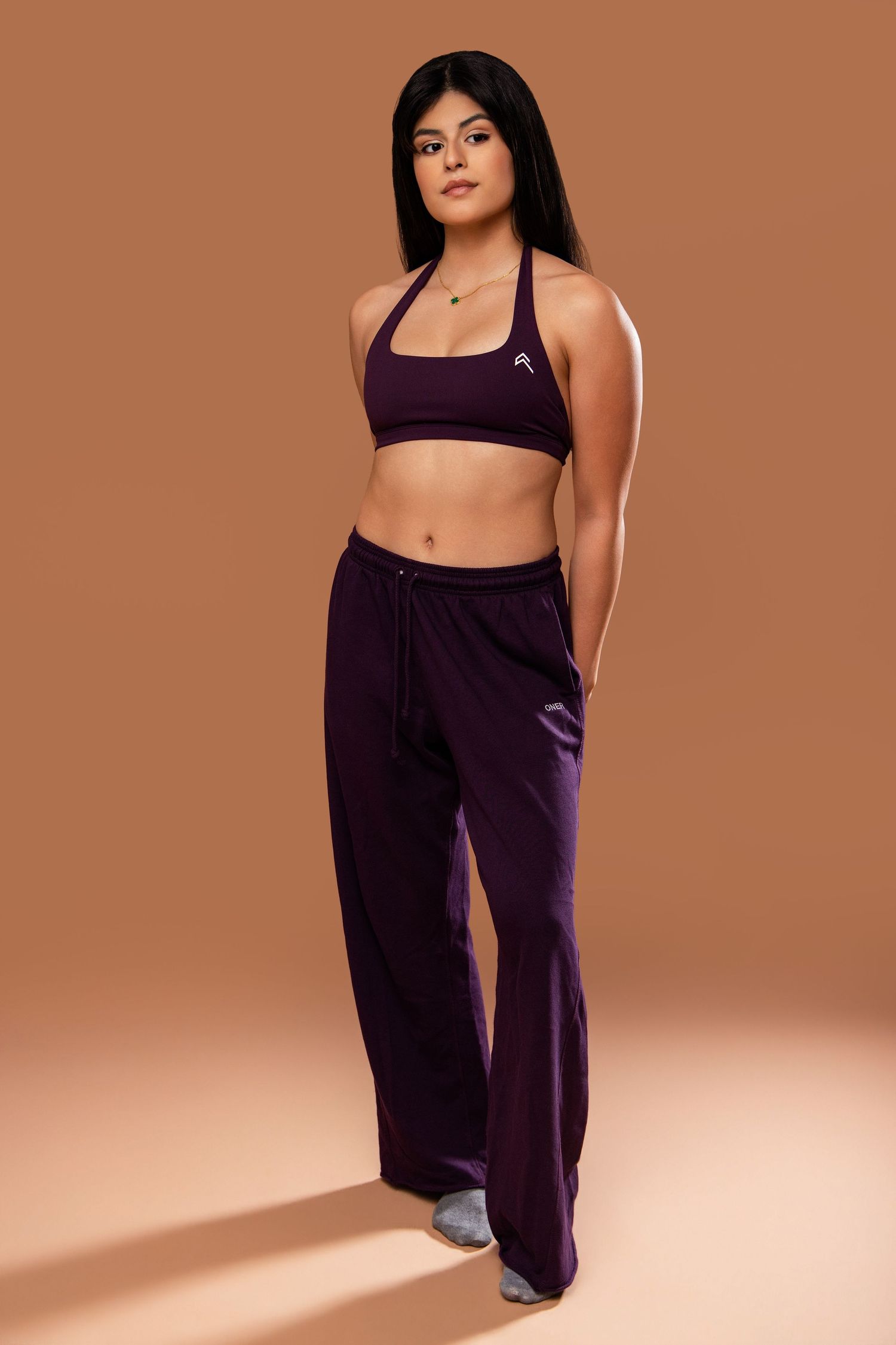 Athletic woman in purple sports bra and matching wide-leg pants poses against warm brown background in fitness photoshoot.