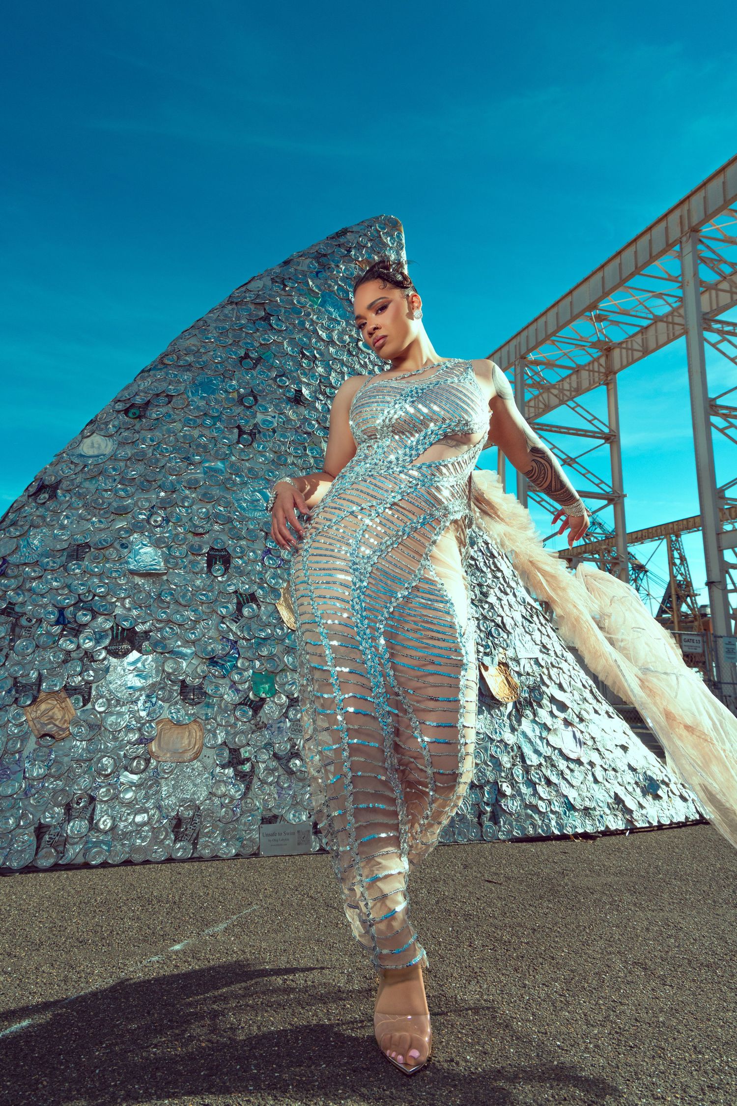A person in a reflective metallic bodysuit poses against a bright blue sky and industrial structure backdrop.
