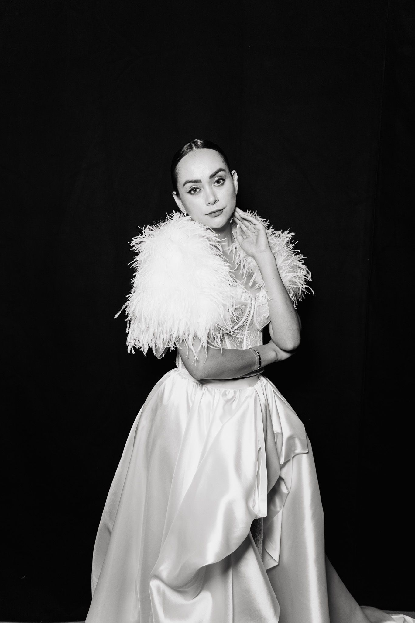 Elegant black and white portrait of a figure in a flowing white satin gown with feather details against dark background.