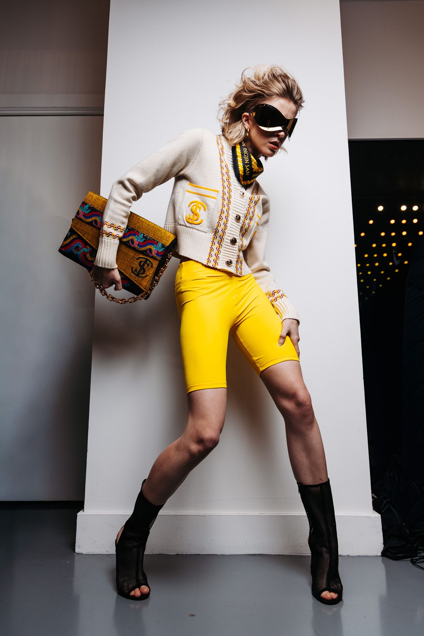 Fashion model in yellow biker shorts and cream blazer poses dynamically with designer handbag and sunglasses.