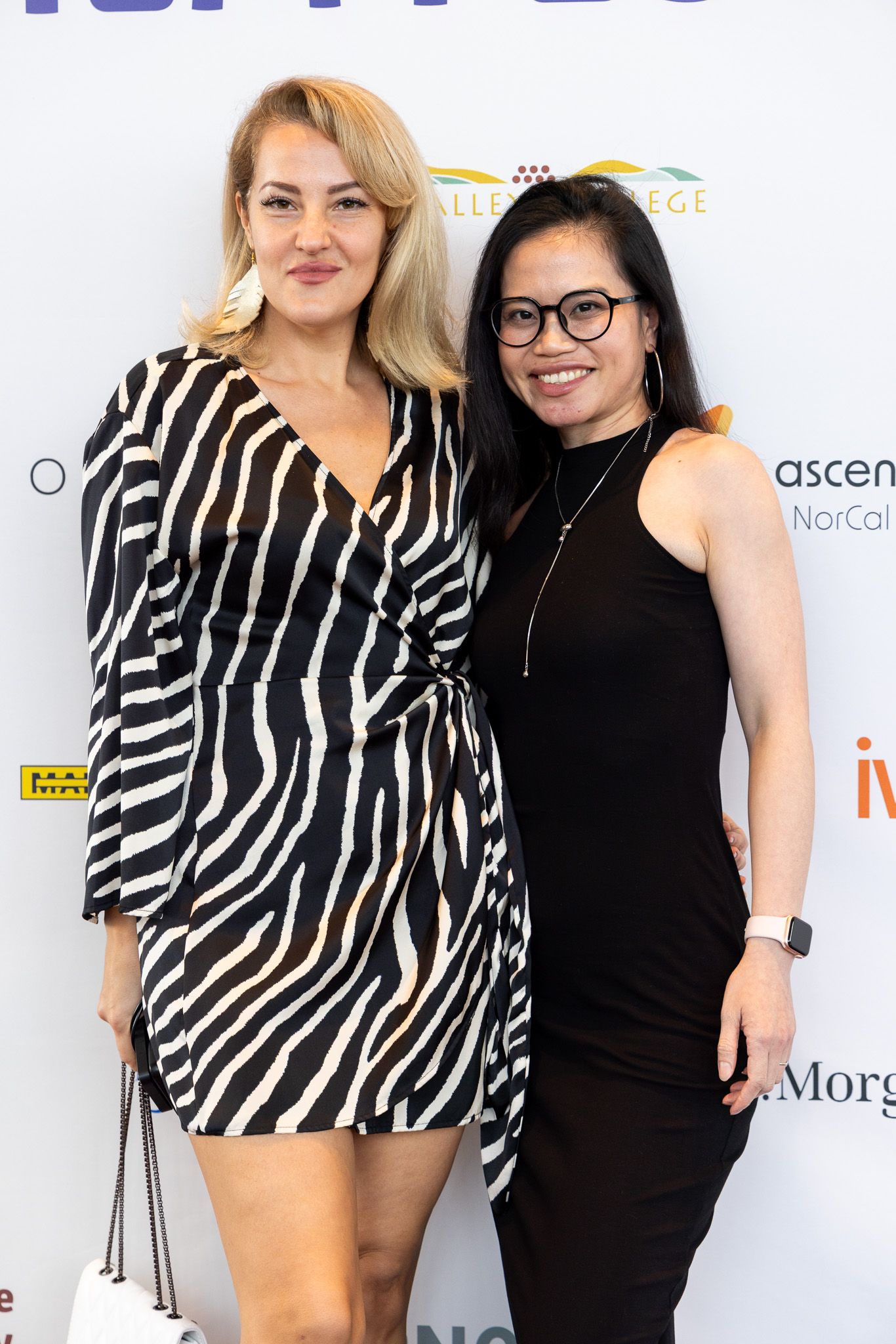 Two people pose together at an event, one wearing a zebra-print top and one in black against a white backdrop with logos.