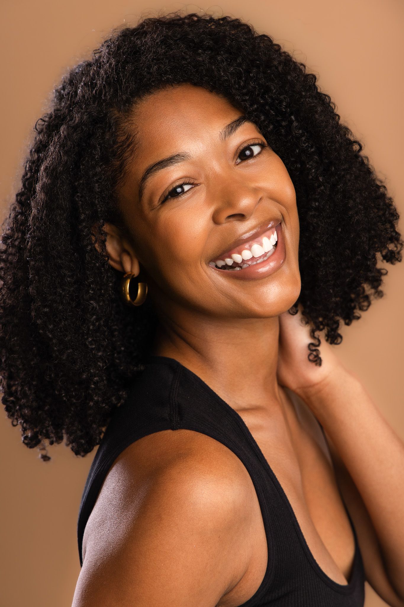 Smiling Black woman with curly hair wearing gold hoop earrings and black top against a warm brown background.