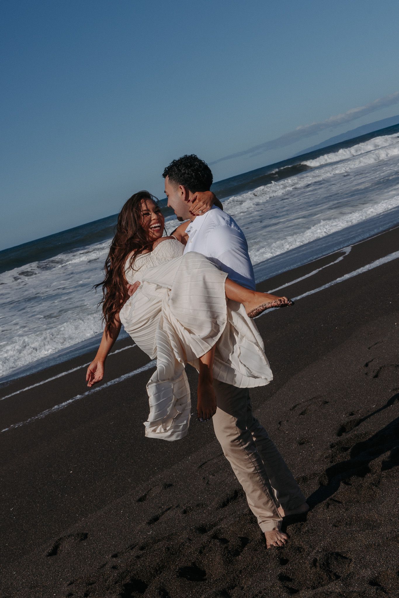 Couple in white outfits dancing romantically on a black sand beach with ocean waves in the background.