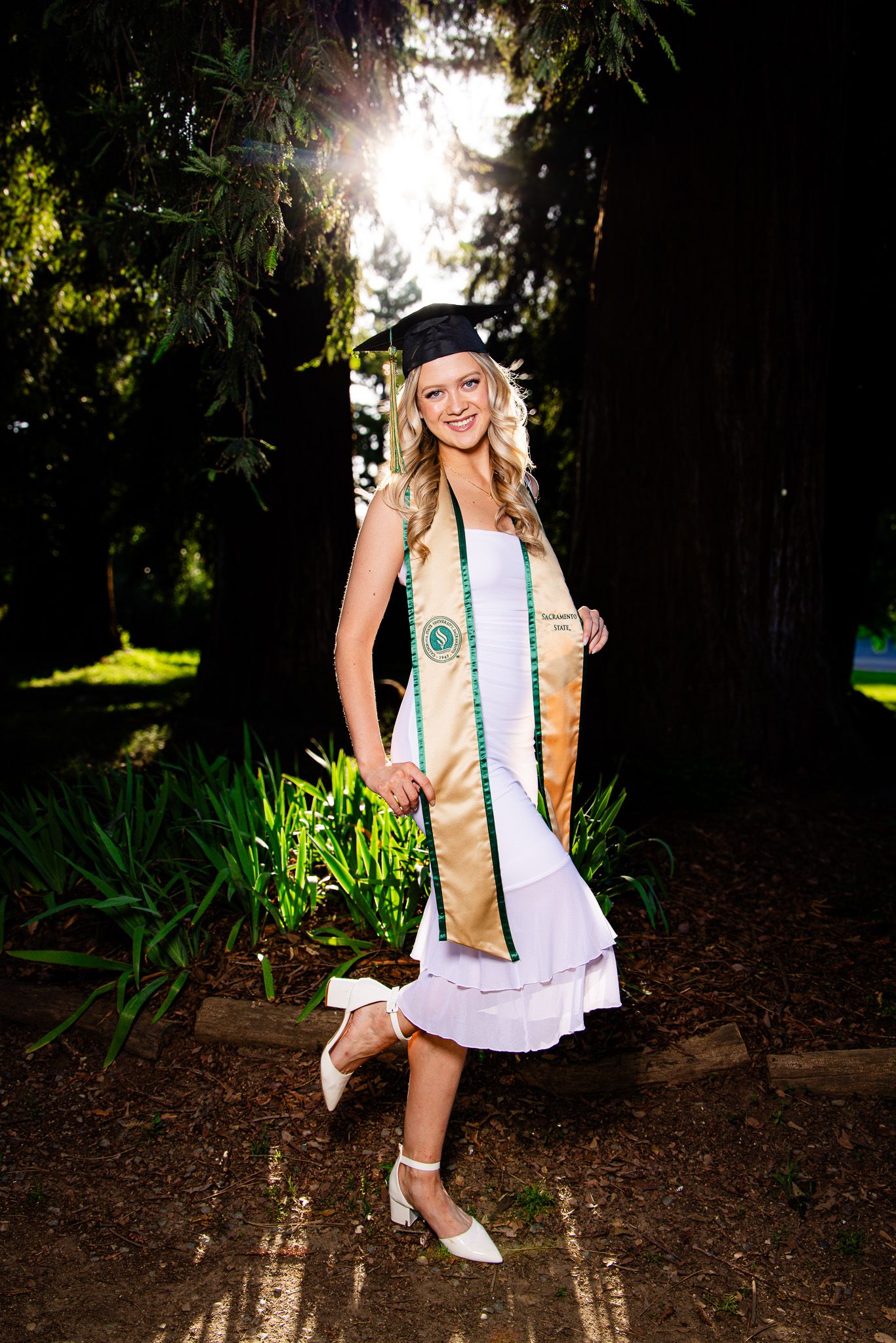 Smiling female graduate in white dress, gold stole, and black cap poses outdoors among tall trees in sunlight.