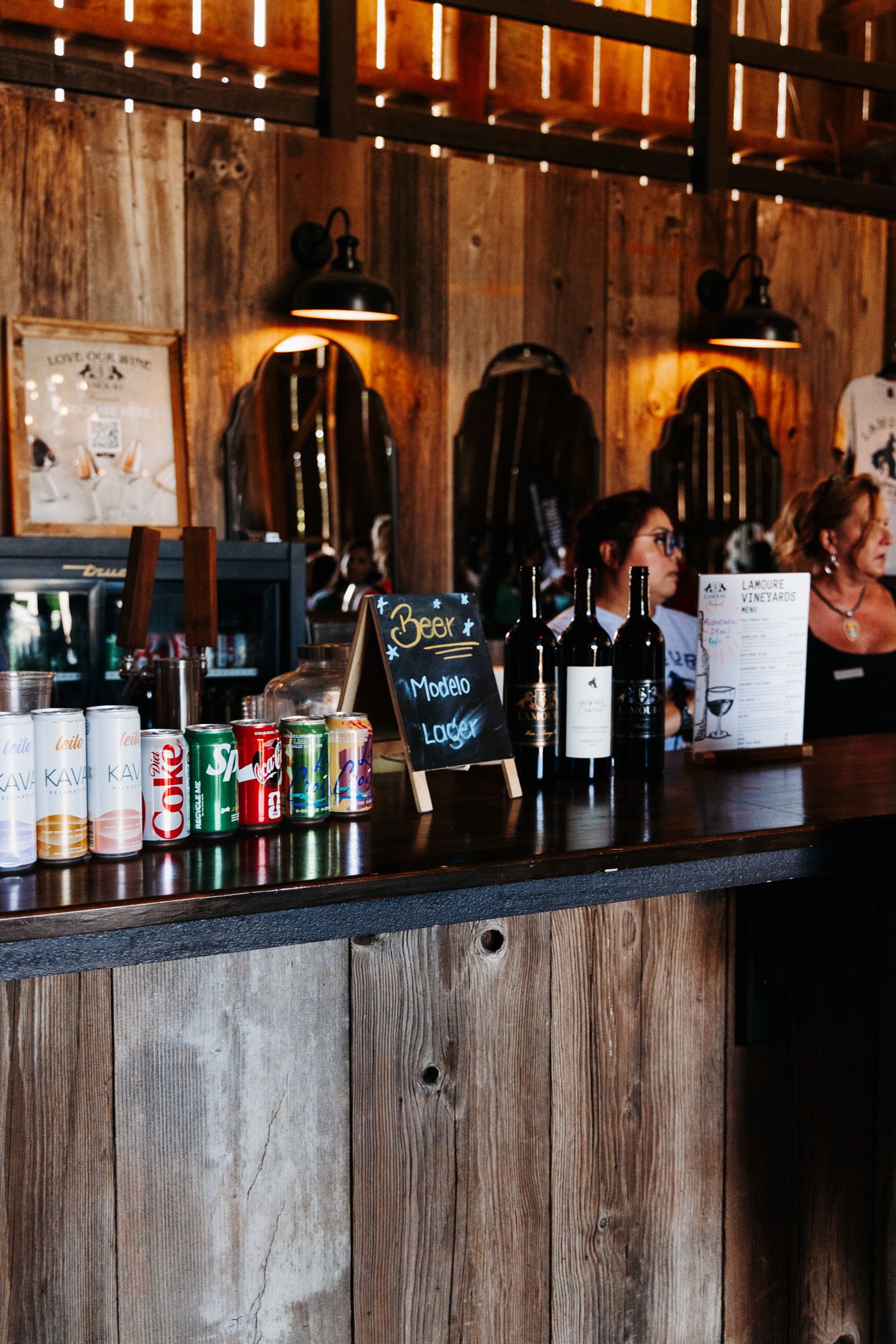 Rustic bar with wine bottles, canned drinks, chalkboard menu, warm string lights, and wooden walls in a cozy venue.