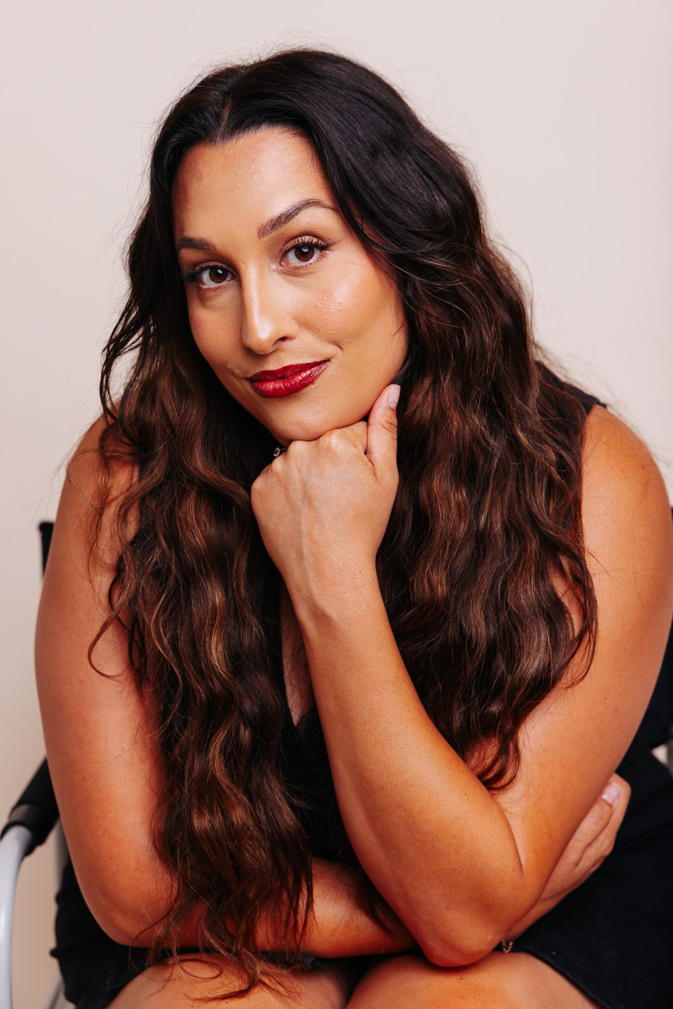 Confident woman with long wavy brown hair and red lips posing with hand under chin.