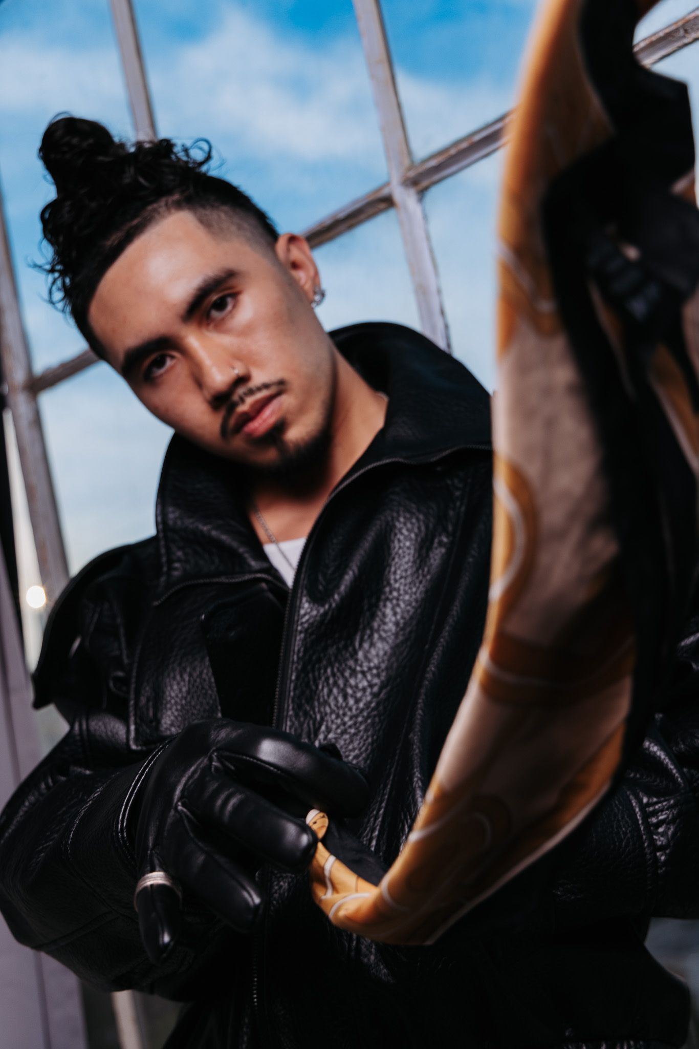 Person in black leather jacket poses against window with dramatic lighting and blue sky background.