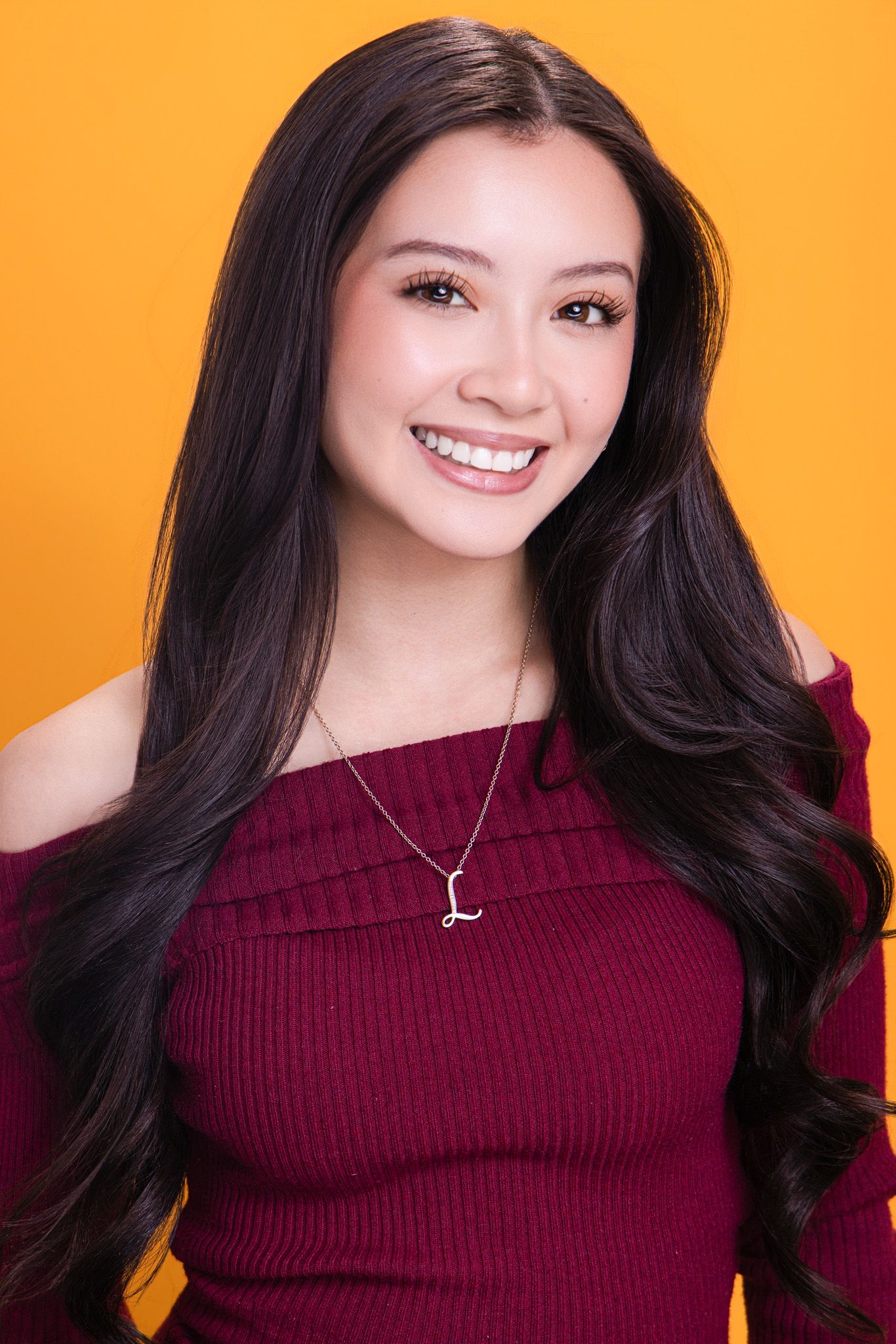 Smiling young woman with long dark hair wearing a burgundy off-shoulder top and initial necklace against yellow background.