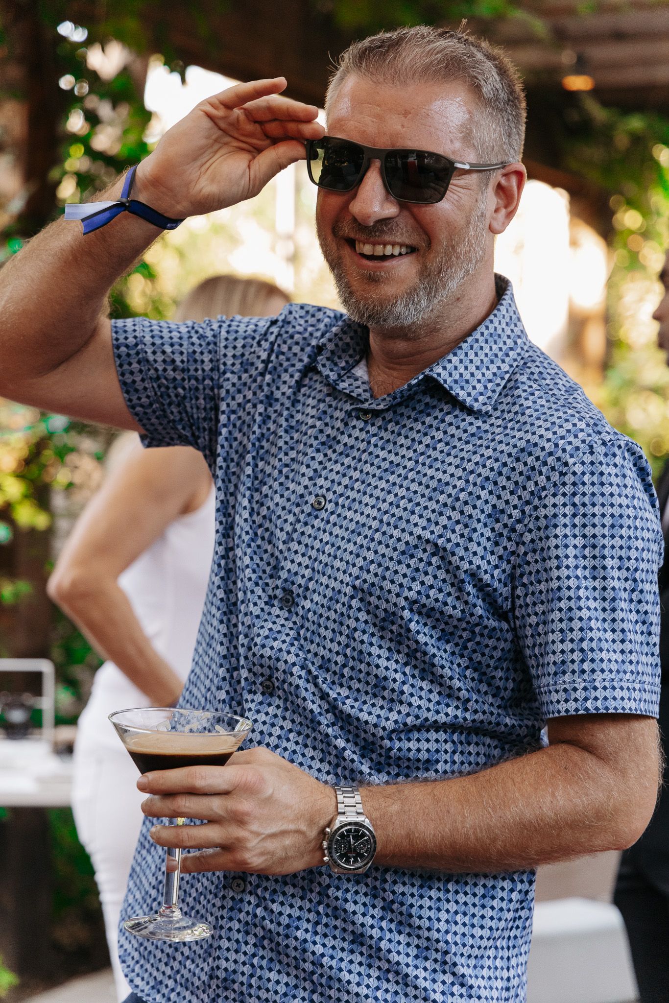 Smiling person in sunglasses and patterned blue shirt holding a cocktail glass at an outdoor event.