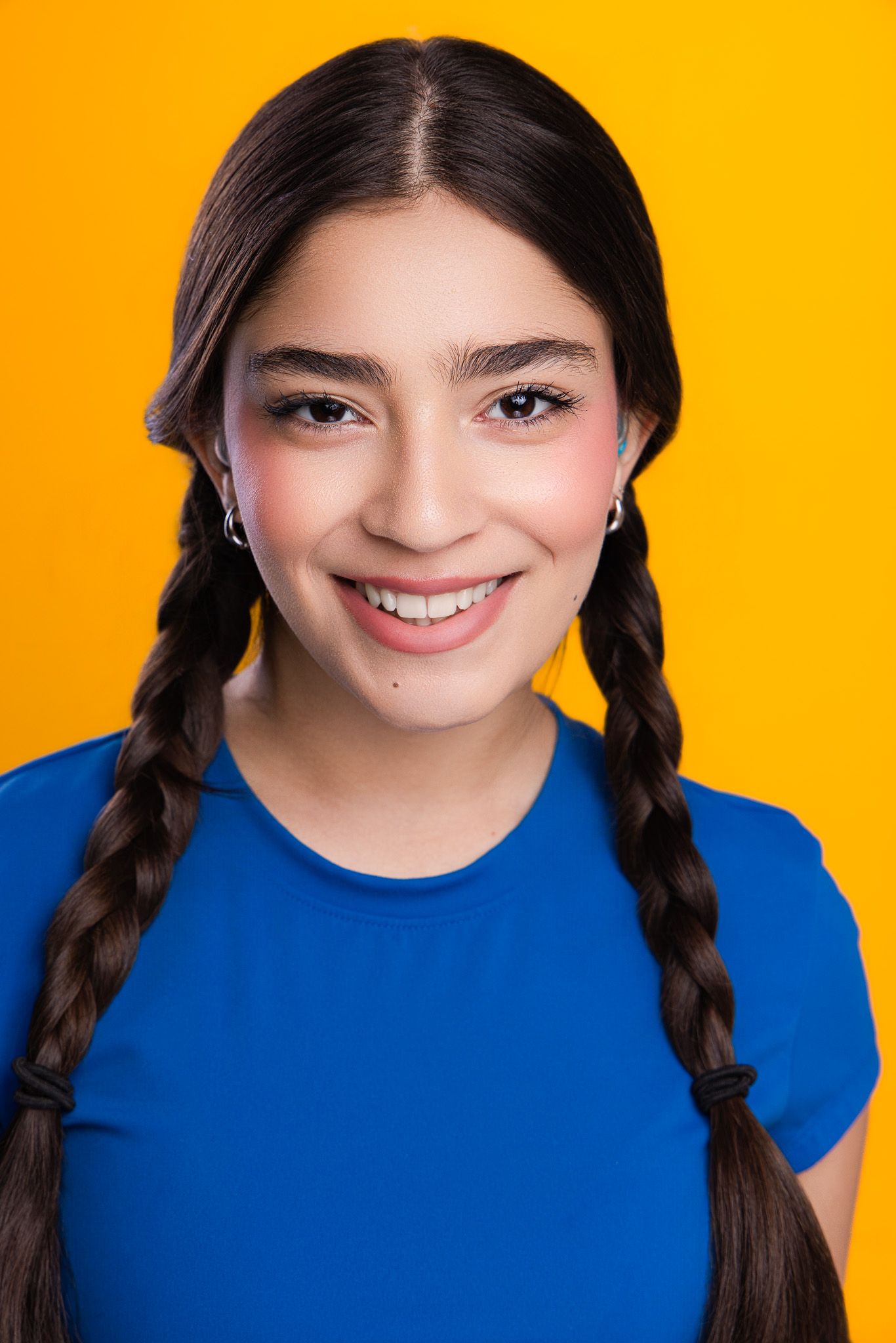 Young woman with dark braided pigtails smiling, wearing a blue t-shirt against a bright yellow background.