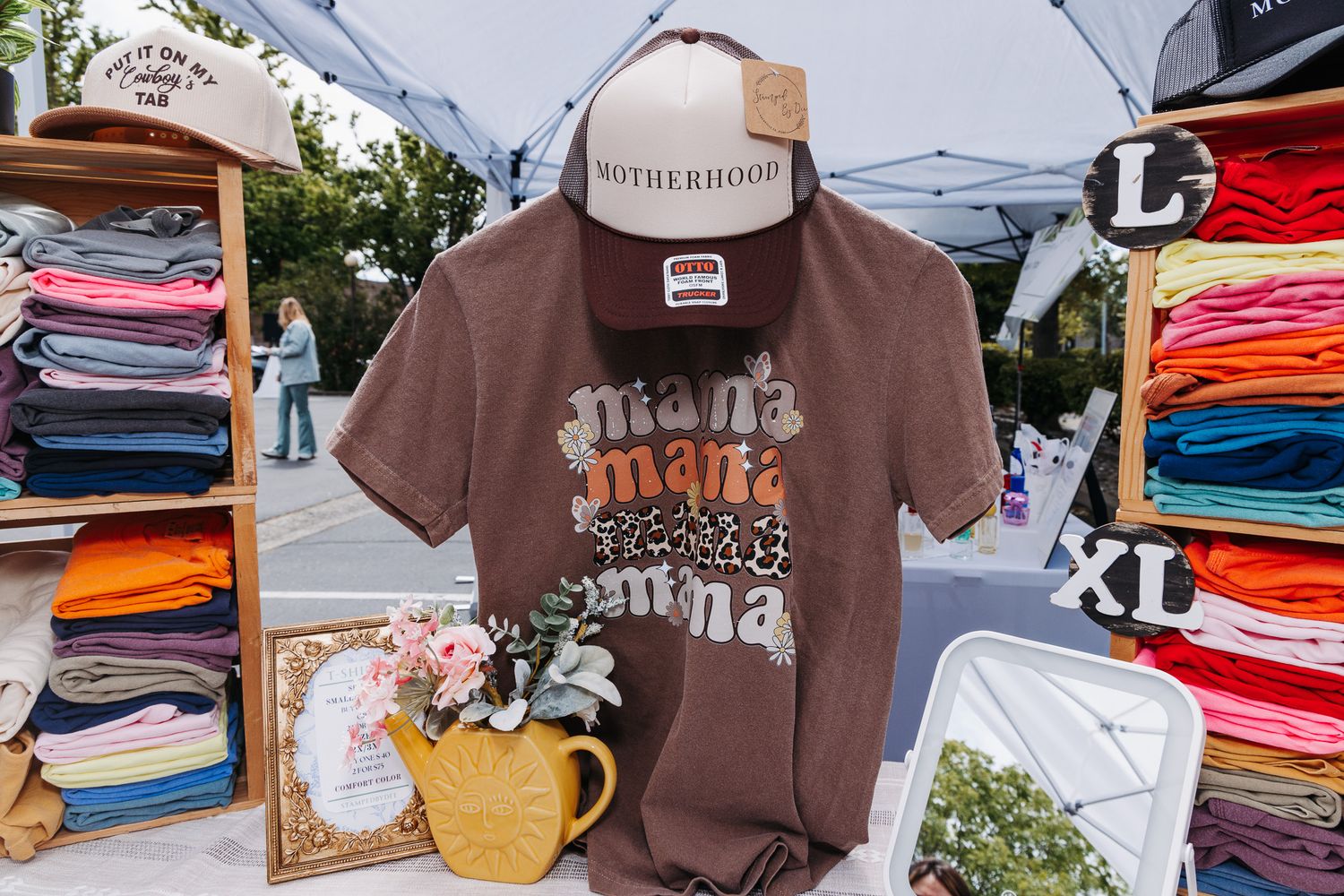 Outdoor market booth displaying brown mama graphic tees and Motherhood trucker hats on a folding table.