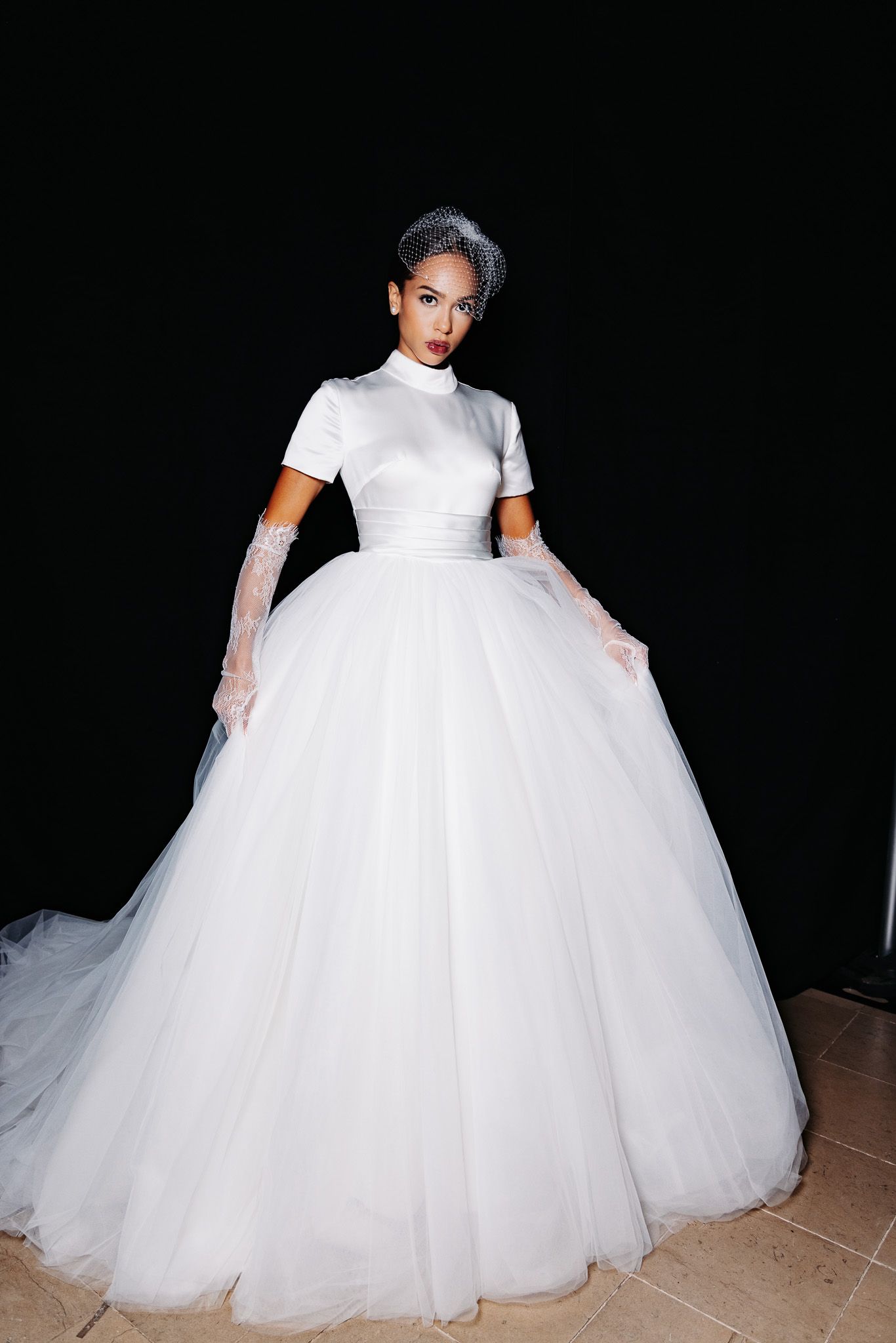 Model showcases a dramatic white ballgown with voluminous tulle skirt on dark background.