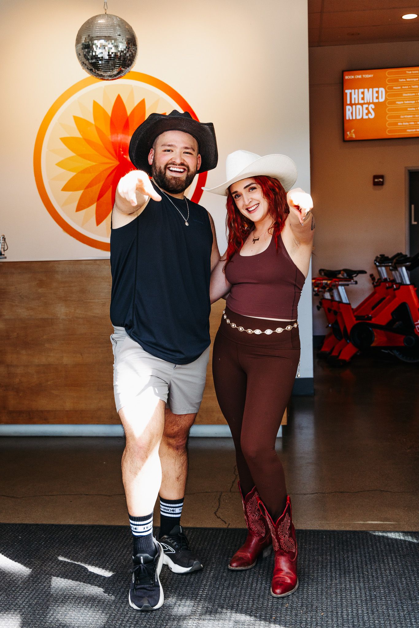 Two people pose energetically in front of a sunflower logo at a fitness studio with a disco ball overhead.