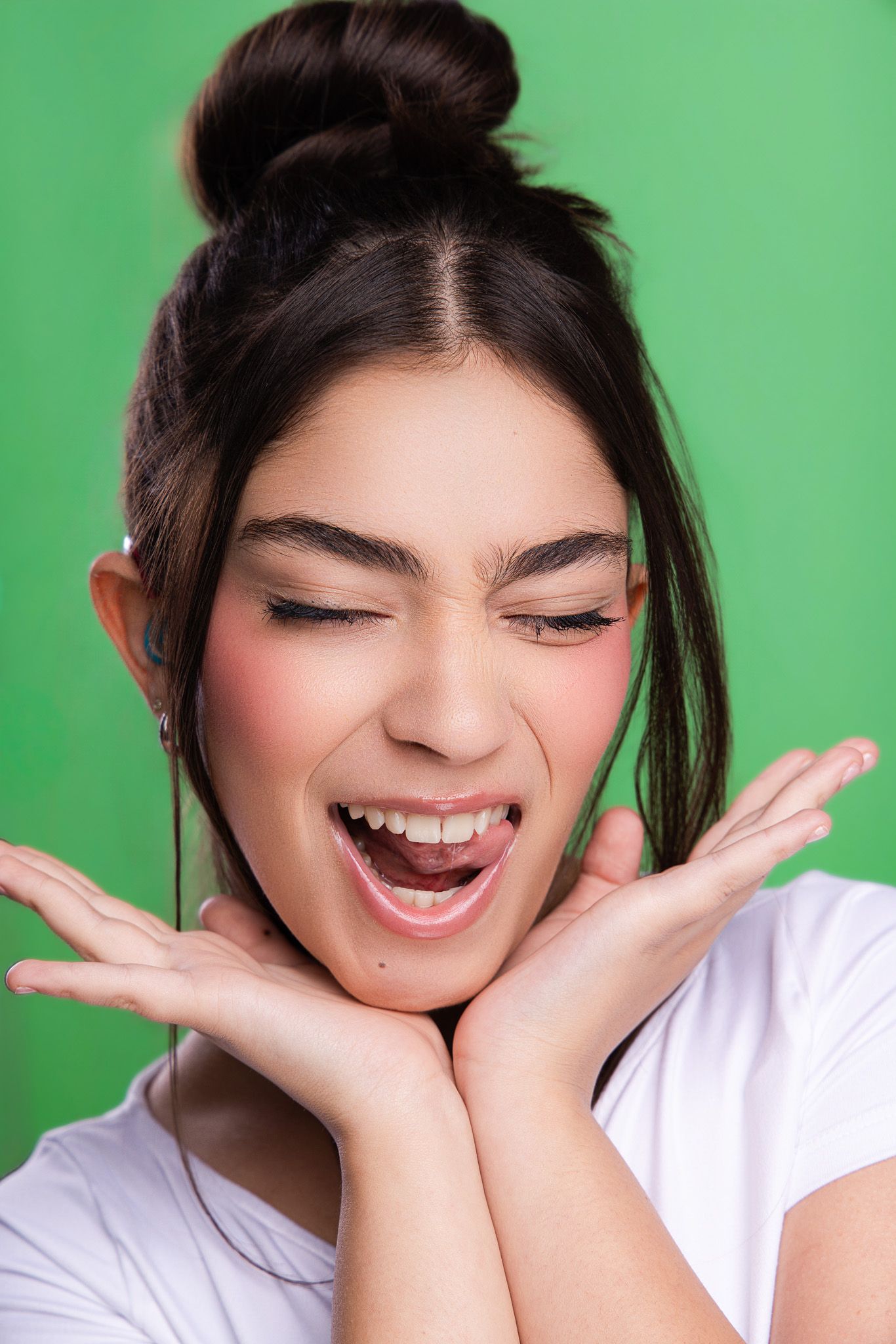 Young woman with hair bun, eyes closed, laughing joyfully with hands framing face against green background.