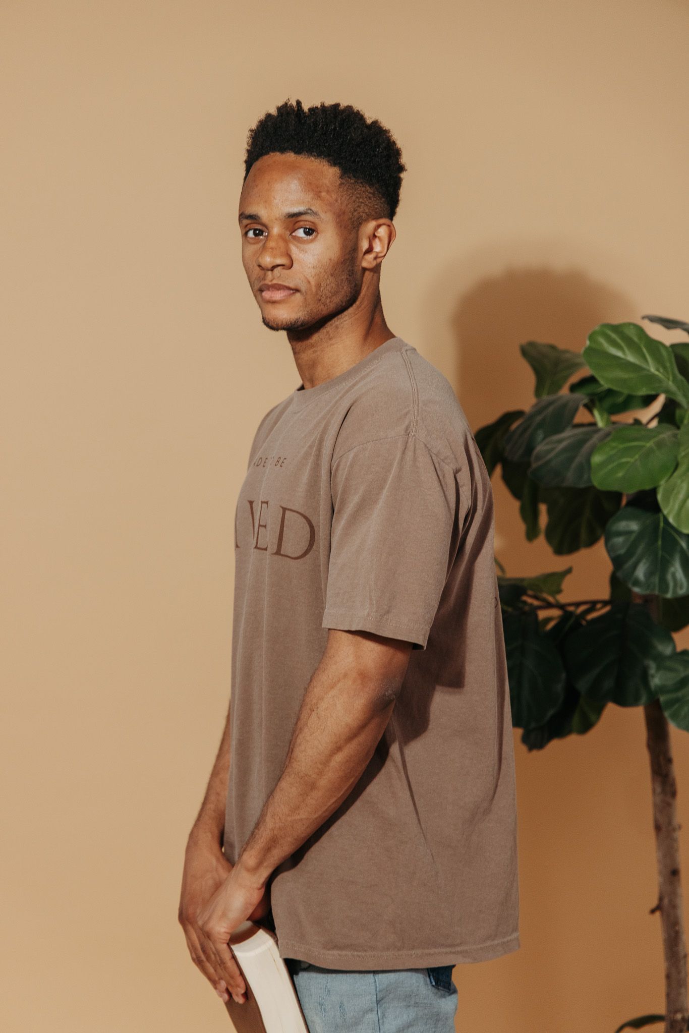 Man in brown t-shirt poses against beige backdrop next to fiddle leaf plant showing product from different angles.