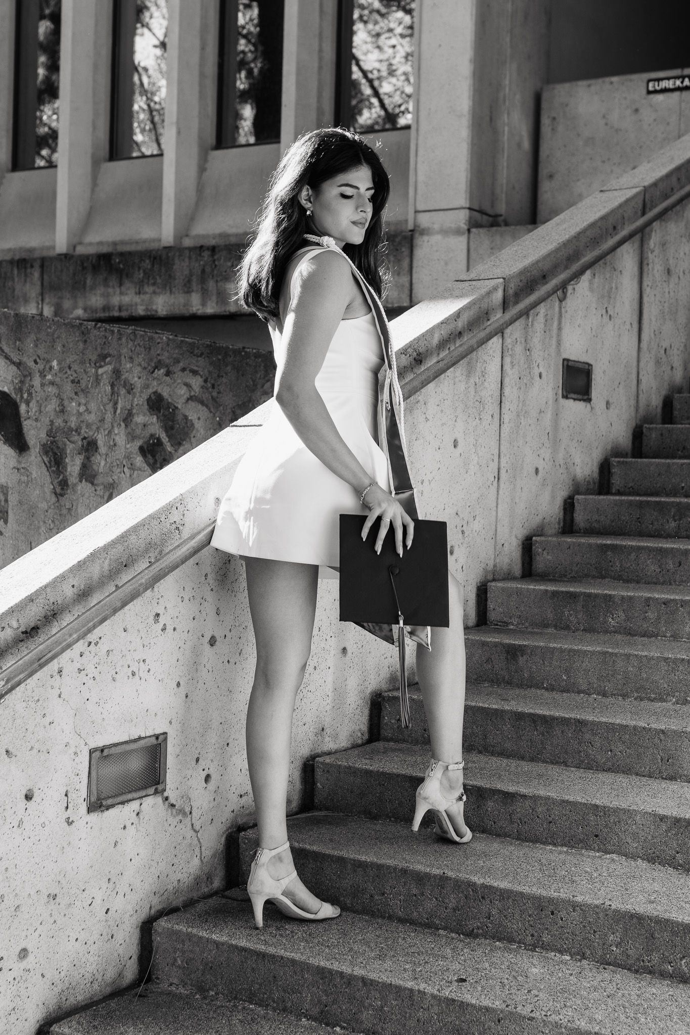 Black and white photo of a person in a white dress and heels posing elegantly on concrete stairs outdoors.