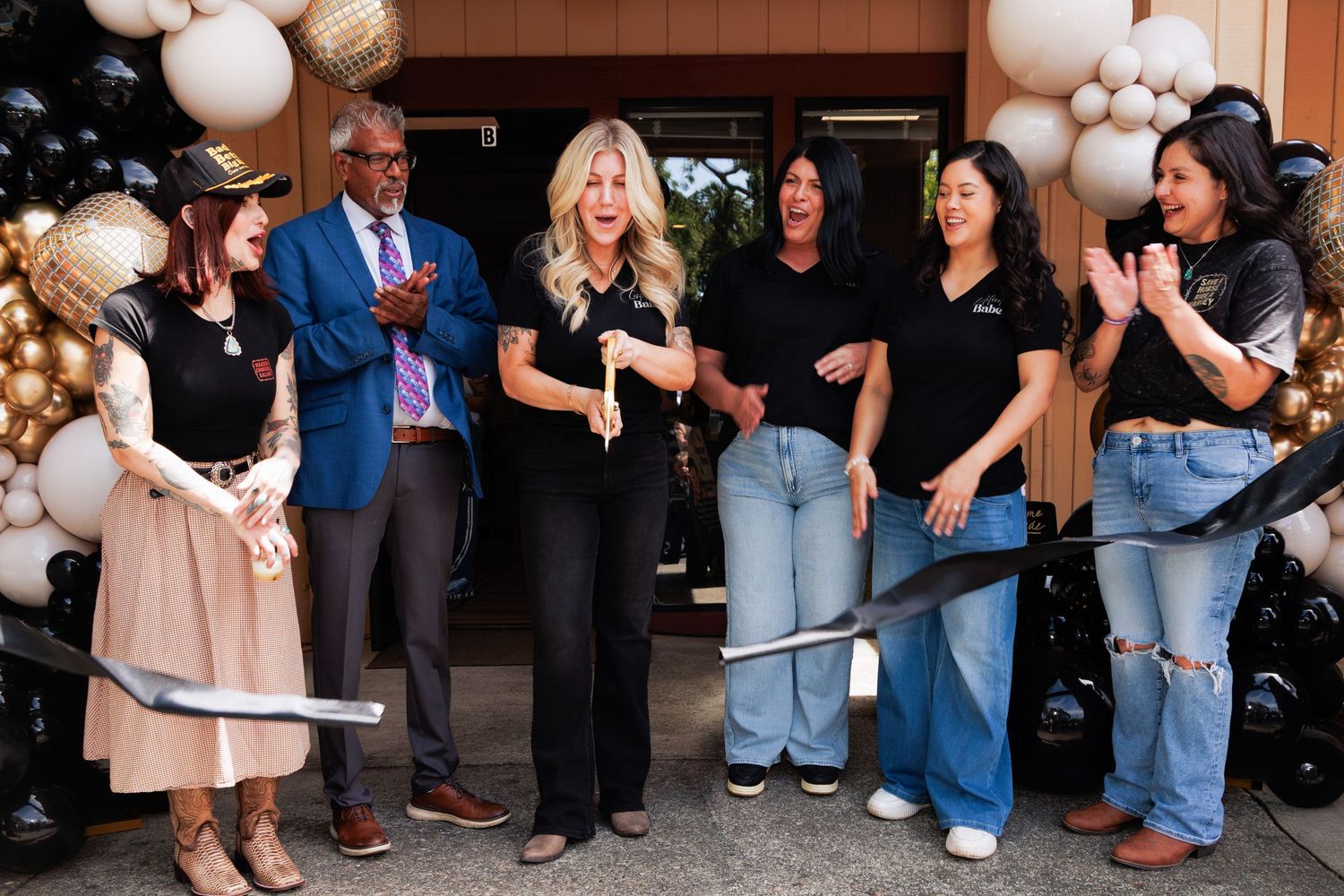 Ribbon cutting ceremony with group of six people surrounded by black and gold balloons.