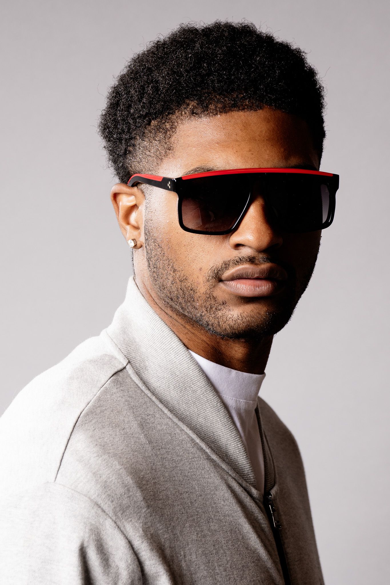 Young Black man with afro hair wearing red-trimmed black sunglasses and gray jacket poses against gray background.