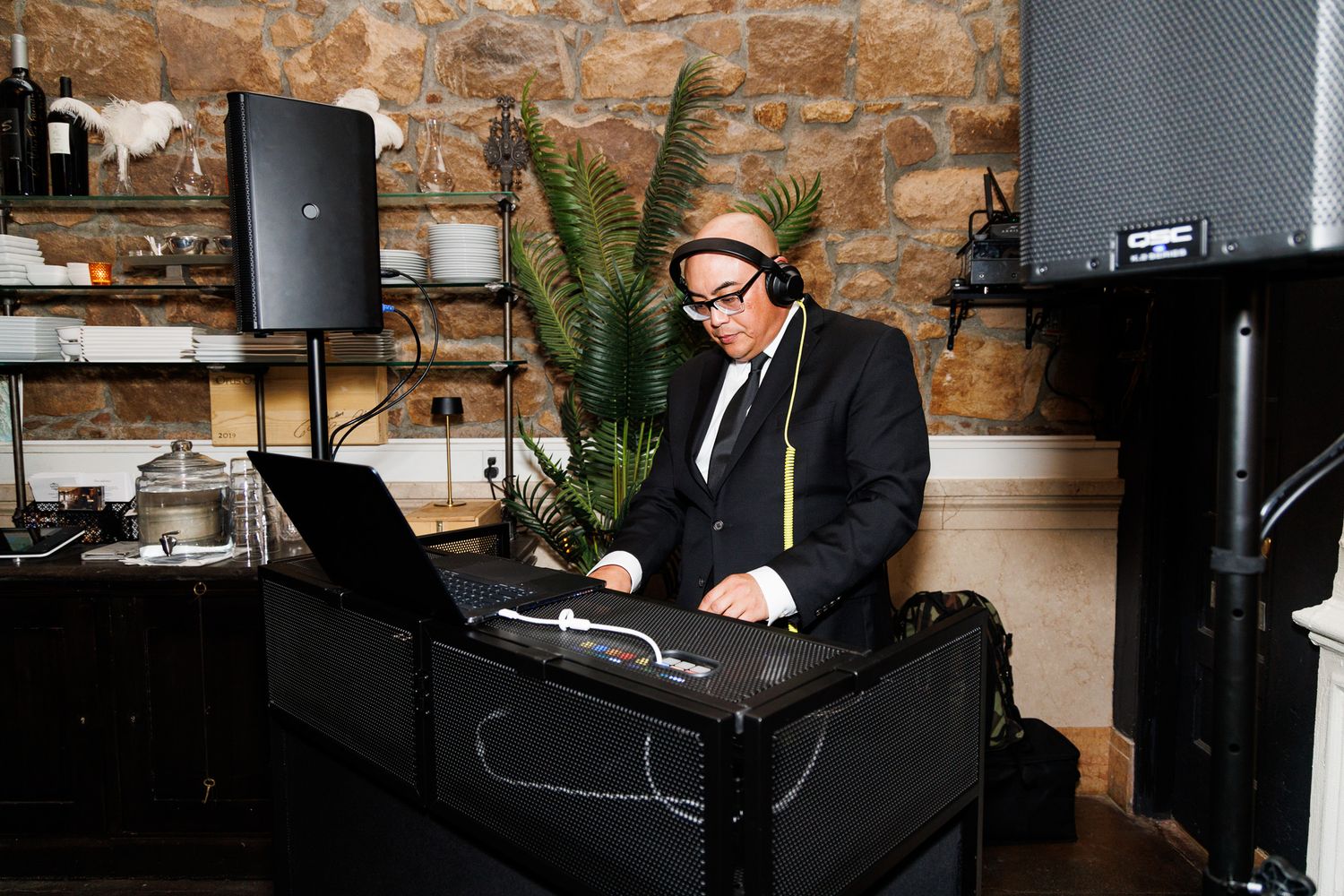 DJ in black suit and hat performs at an upscale indoor event using laptop and mixer.