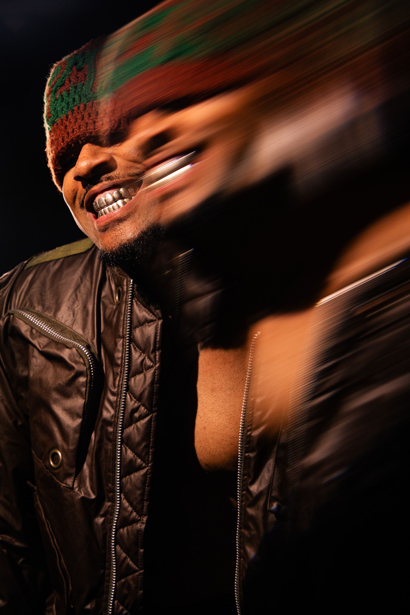 Smiling man in quilted black jacket and multicolor beanie with motion blur effect in dark studio.