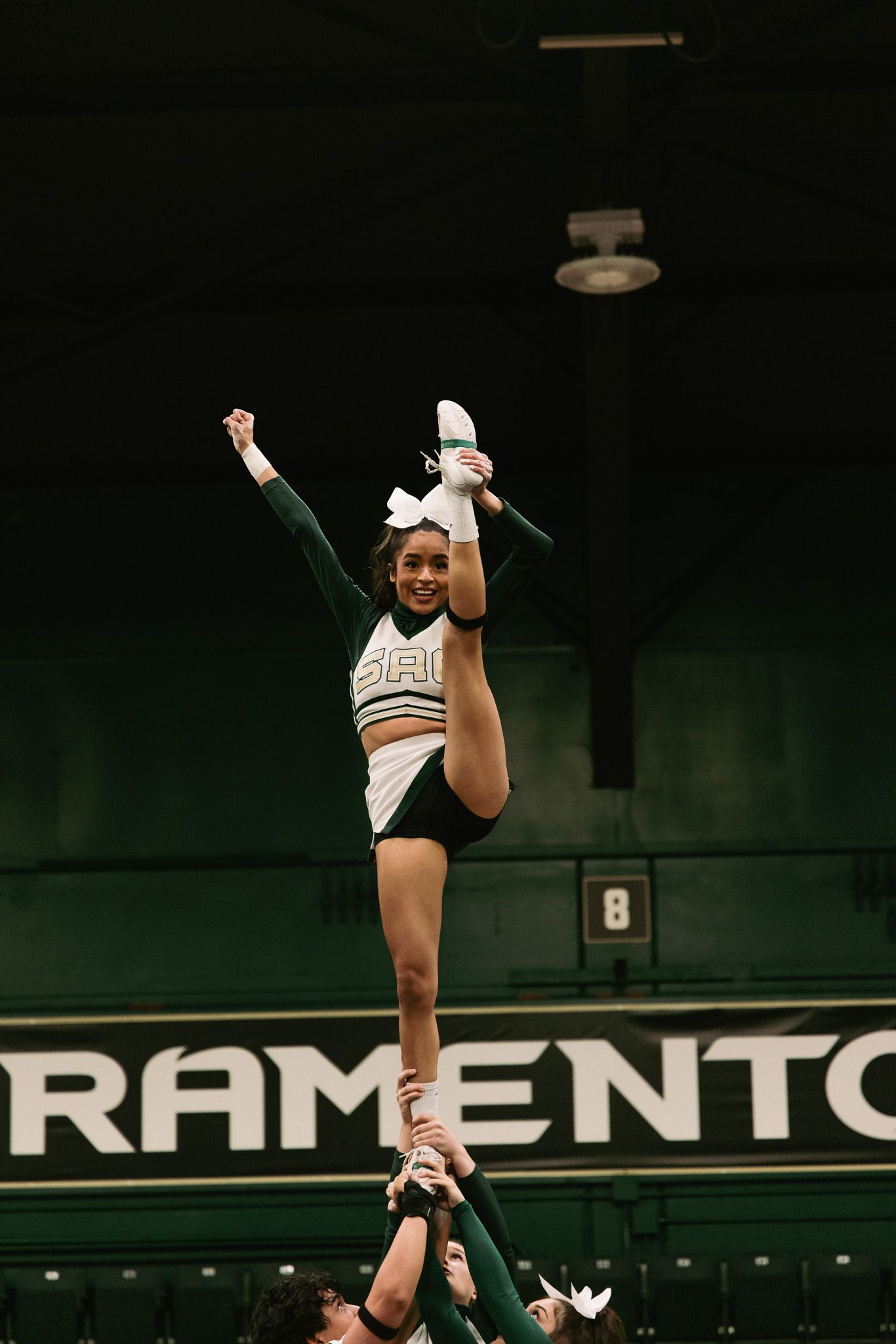 Sacramento cheerleader performs a high heel stretch stunt, lifted by teammates in a green and white uniform indoors.