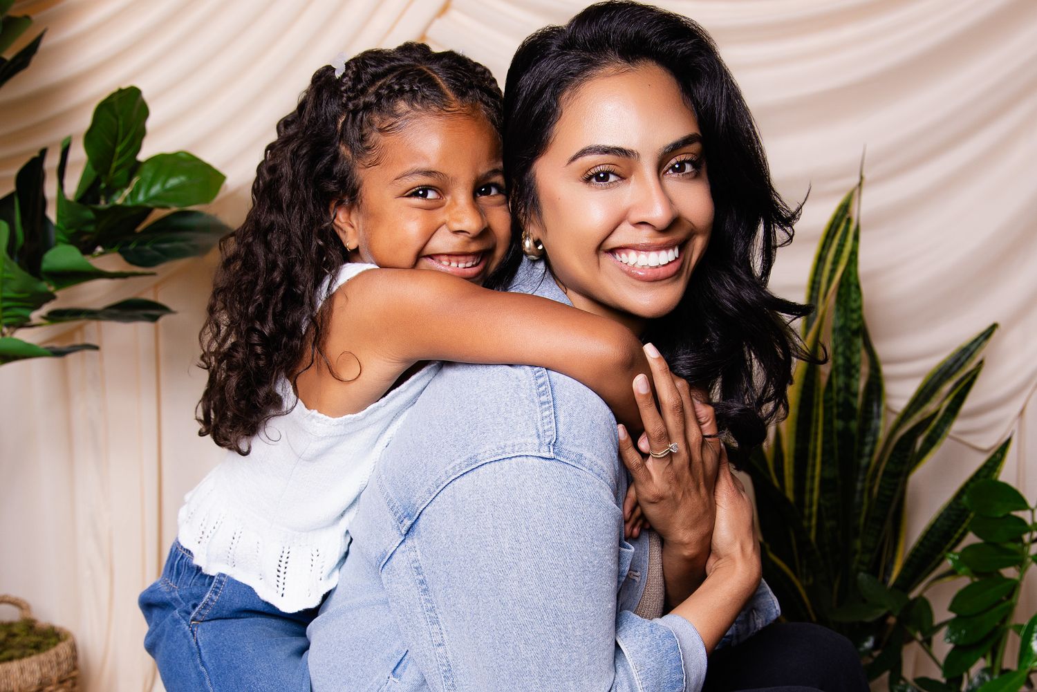 A young girl hugs a smiling woman from behind, both wearing jeans, surrounded by green plants.