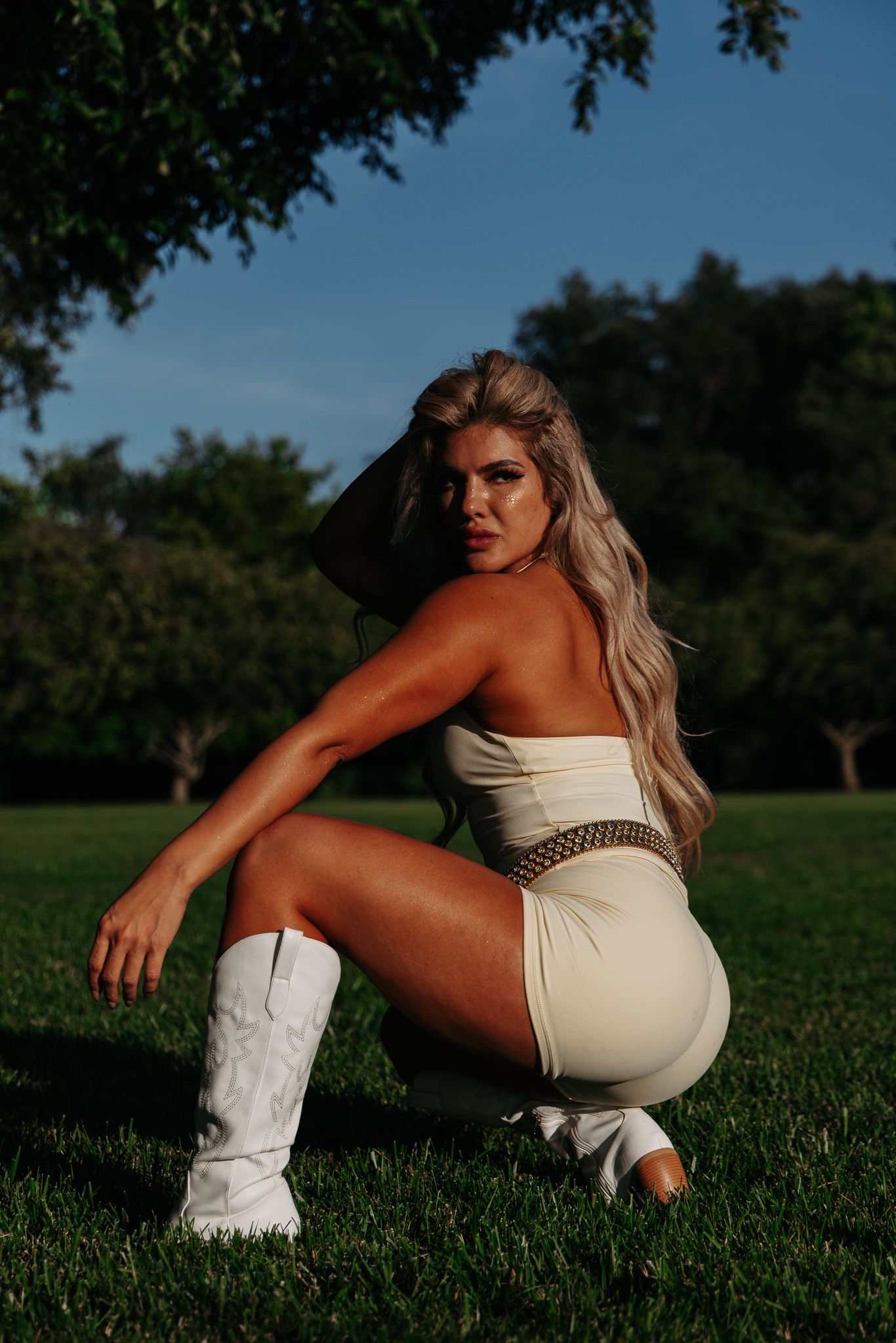 Blonde woman in white strapless bodysuit and cowboy boots posing on green grass with trees in background.