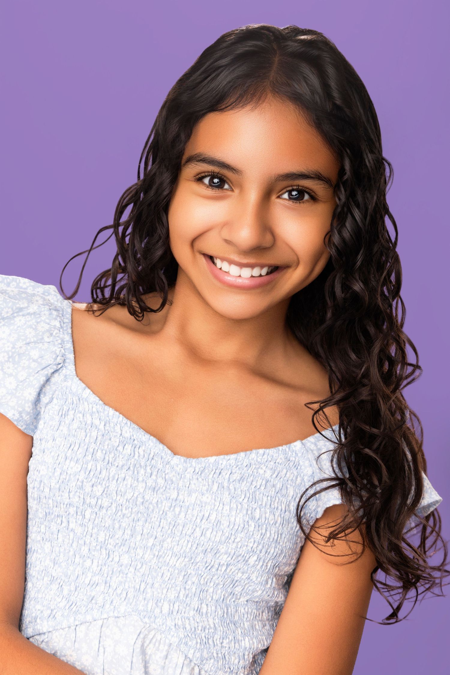 Portrait against purple background wearing white top with natural curly dark hair and bright smile.