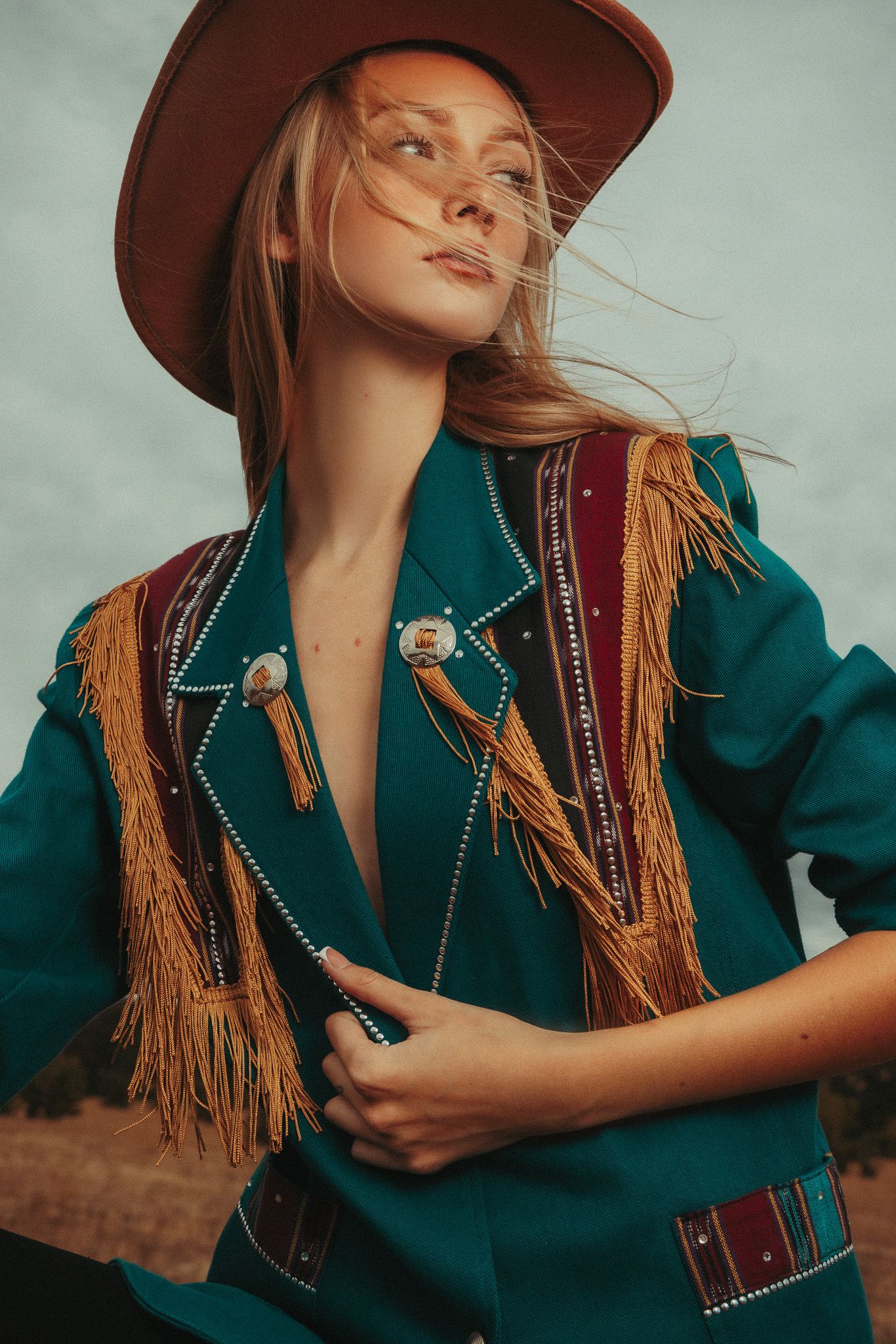 Stylish western-inspired outfit featuring a teal fringe jacket with brown hat against moody sky background.