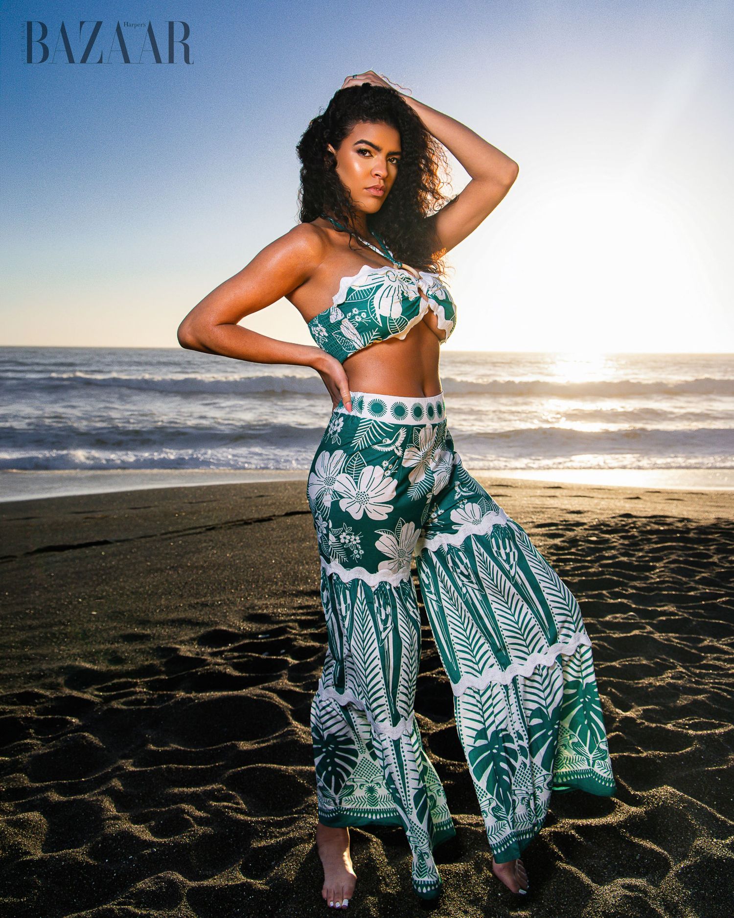 Model poses on beach at sunset wearing green tropical print crop top and wide-leg pants for Harper's Bazaar.