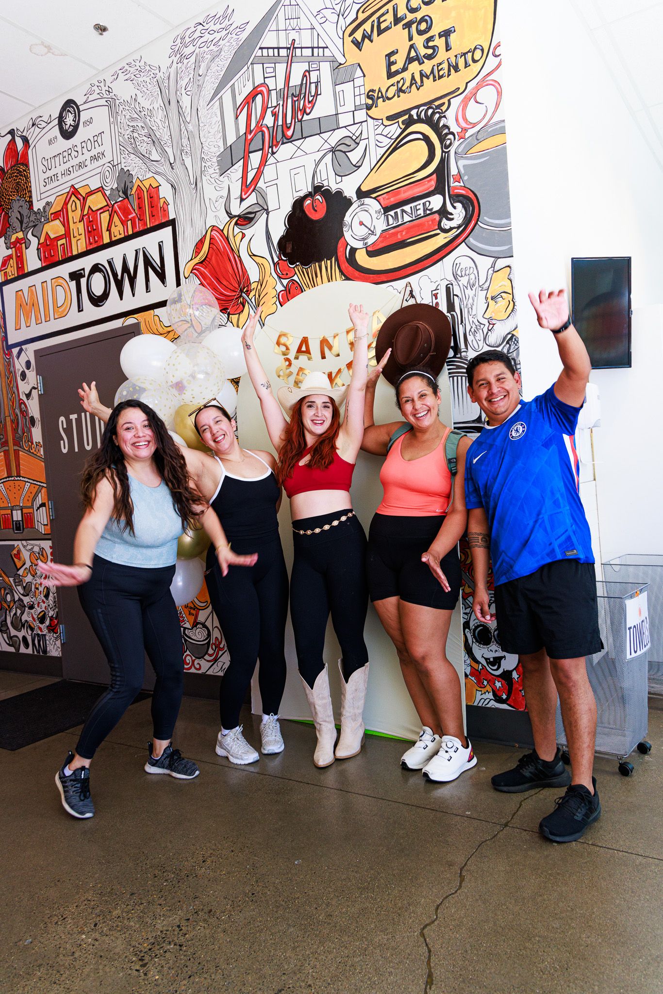 Five people pose joyfully in front of a colorful East Sacramento and Midtown mural, raising arms in celebration.