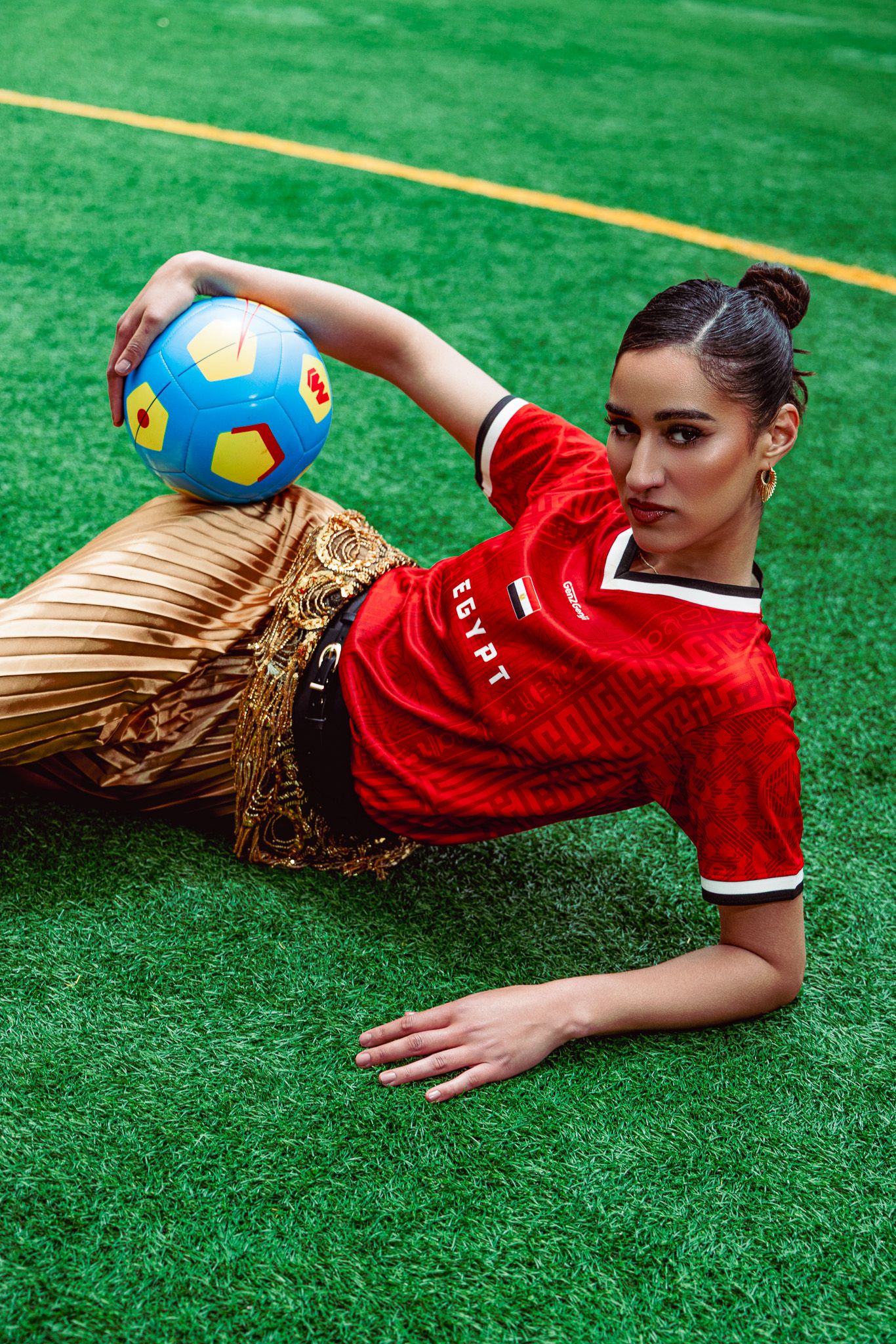 Woman in red Egypt jersey lying on green turf holding a blue and yellow soccer ball wearing a traditional skirt.