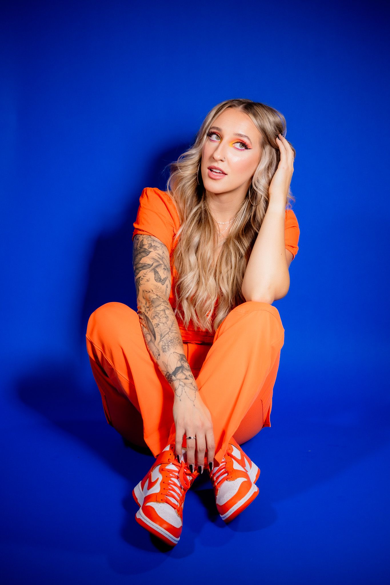 Model in bright orange jumpsuit and matching sneakers poses against vivid blue backdrop in artistic fashion series.