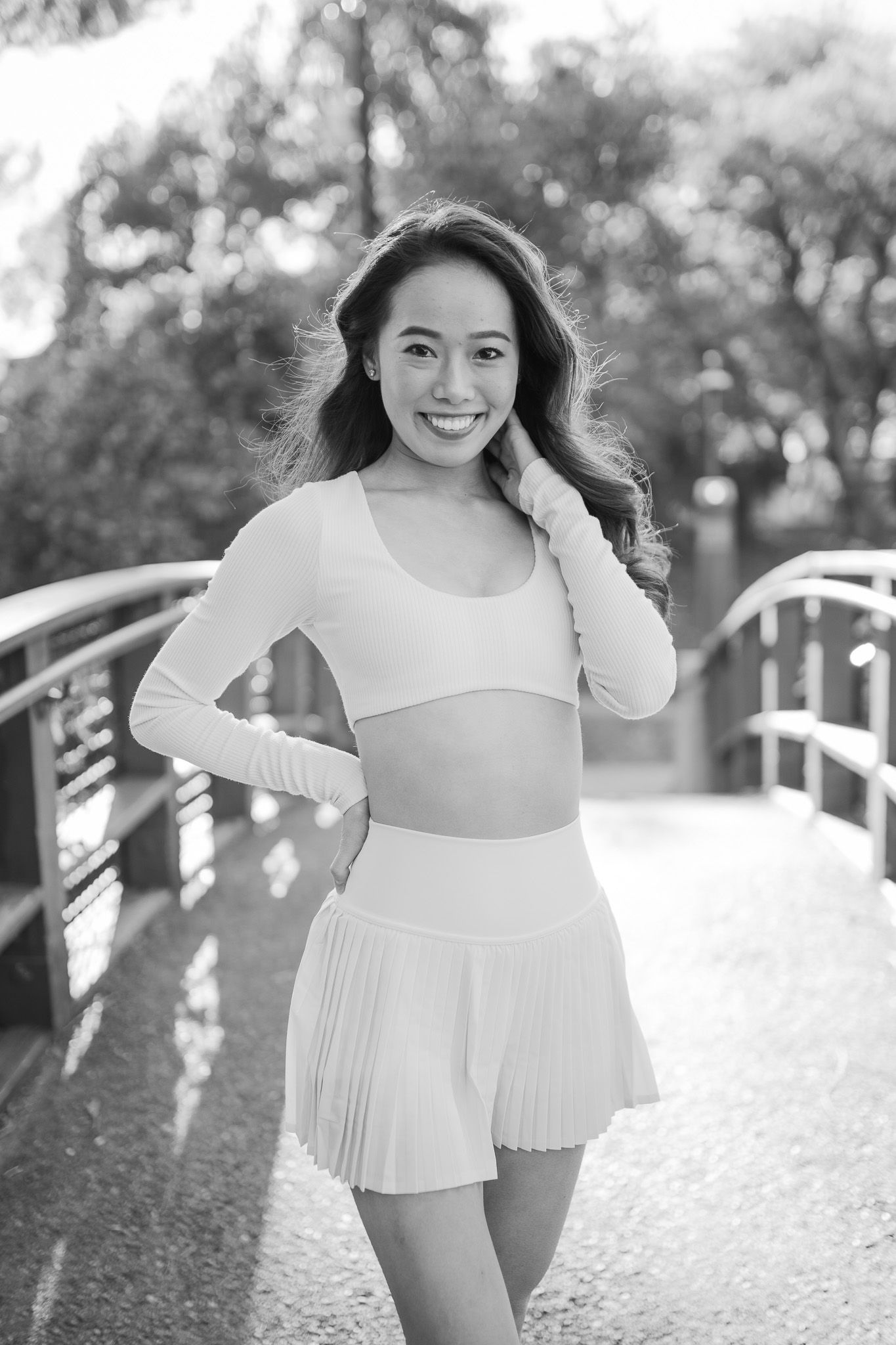 Black and white photo of a person in a white dance outfit with pleated skirt posing on a bridge in a park setting.