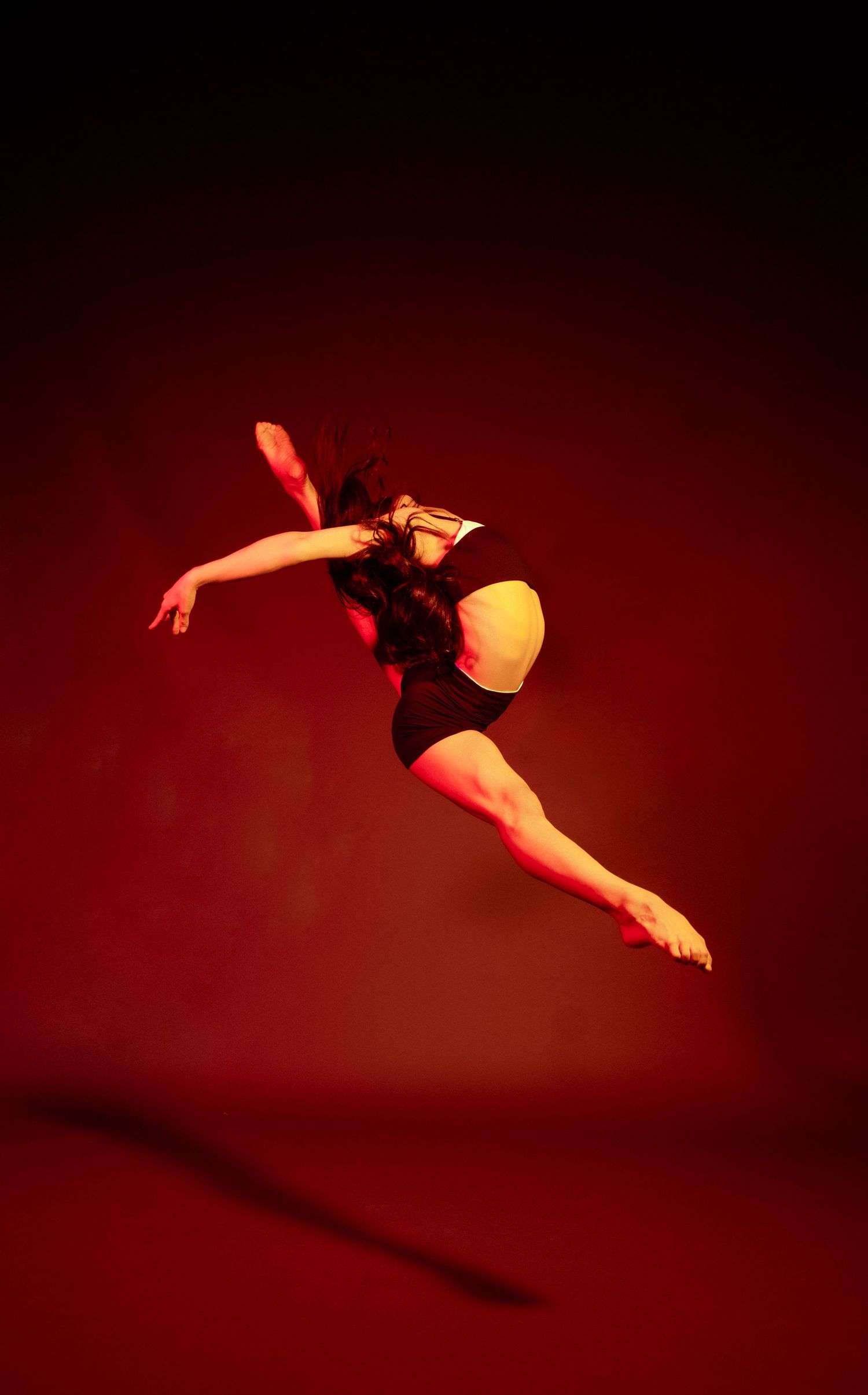 Graceful dancer leaping through air in elegant pose against deep red background.