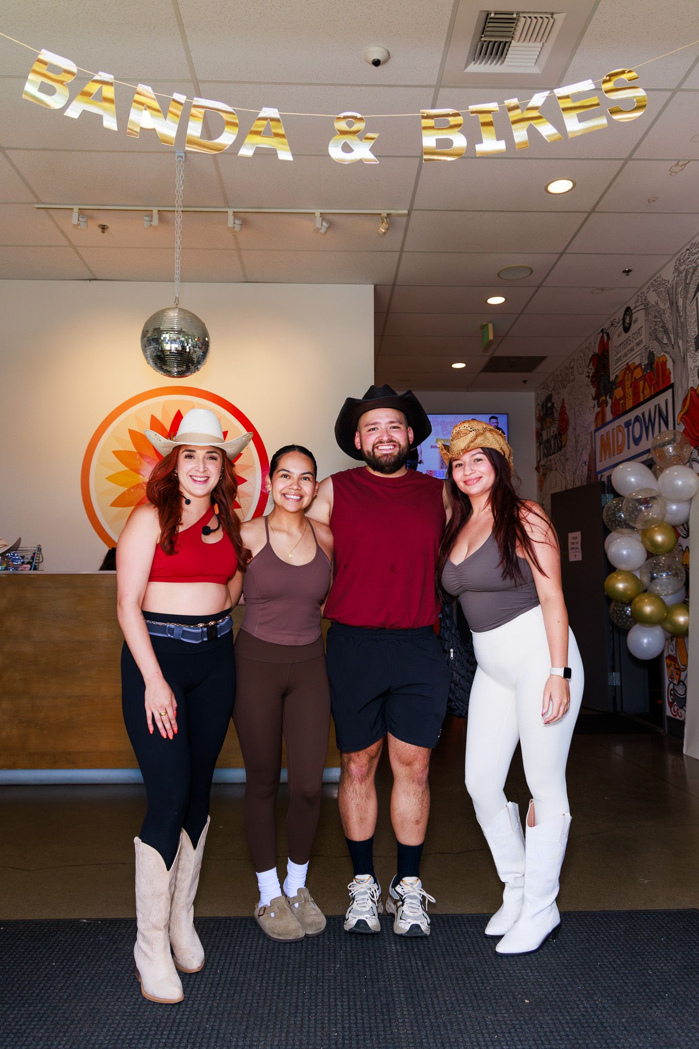 Four friends pose together at a Banda & Bikes event, dressed in western-themed outfits inside a fitness studio.