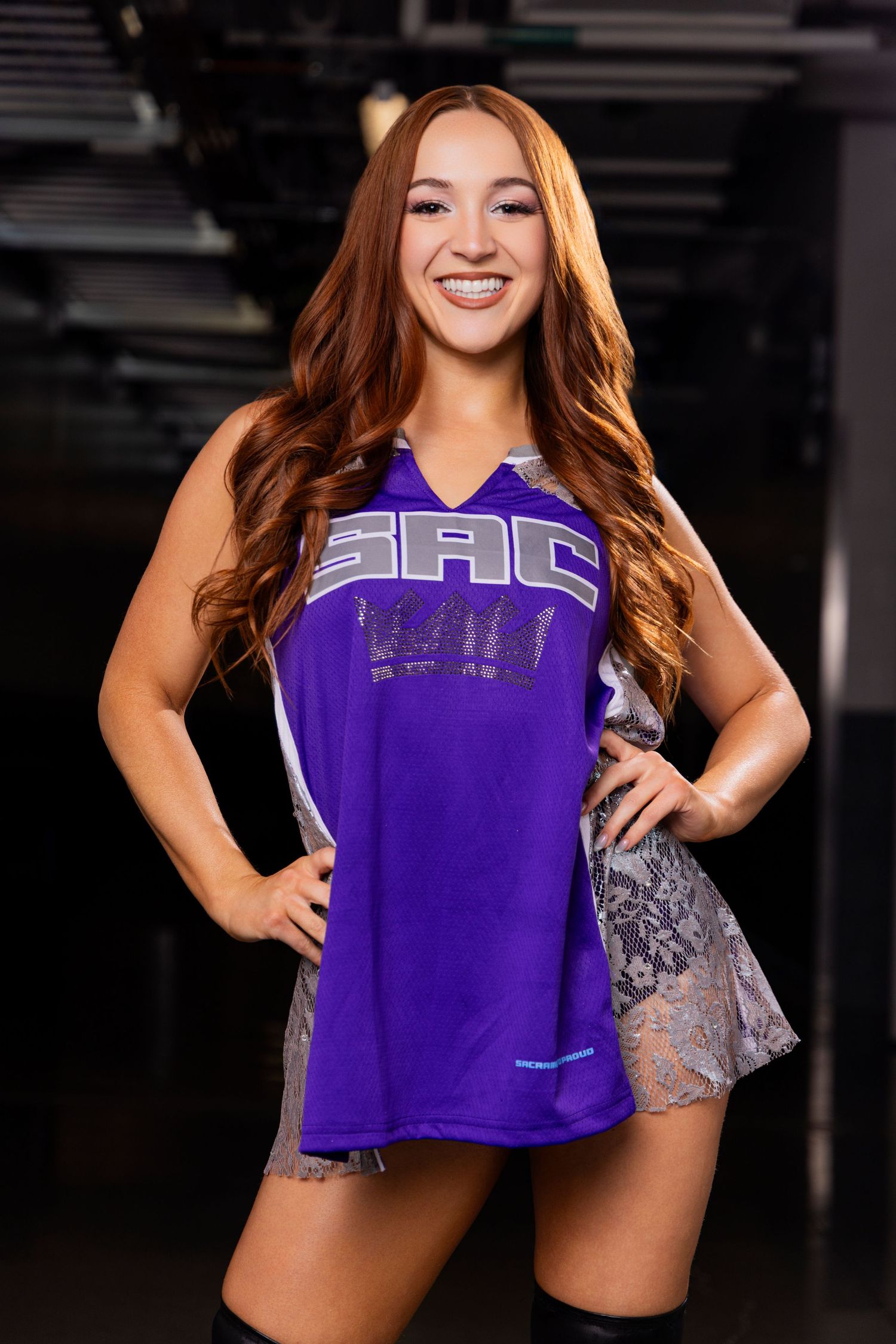 Smiling woman in purple SAC jersey dress with long auburn hair posing with hands on hips in arena.