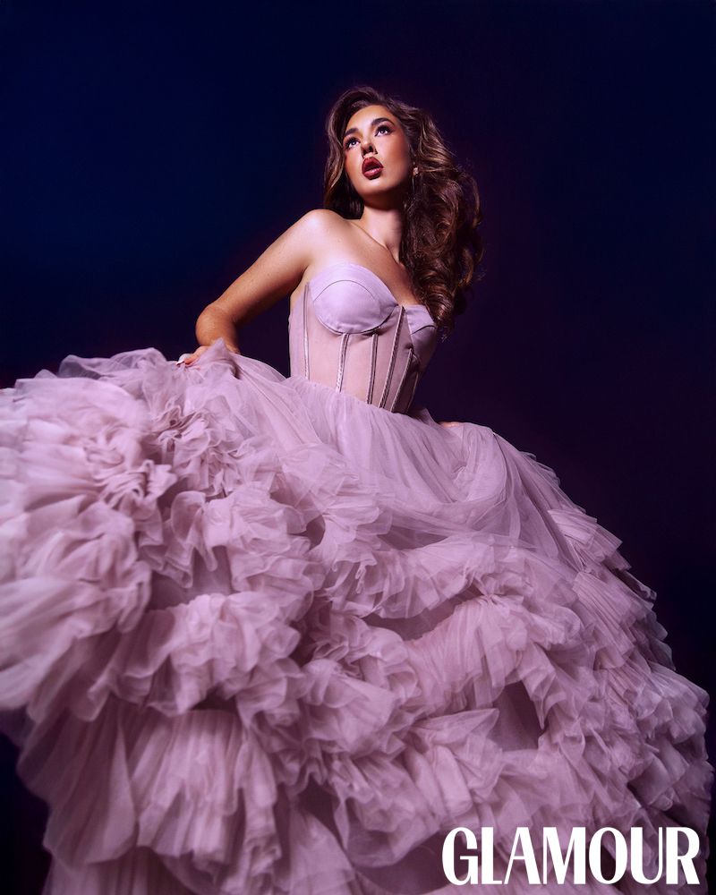 A model poses in a flowing pink tulle gown against a dark backdrop for a glamorous fashion photoshoot.
