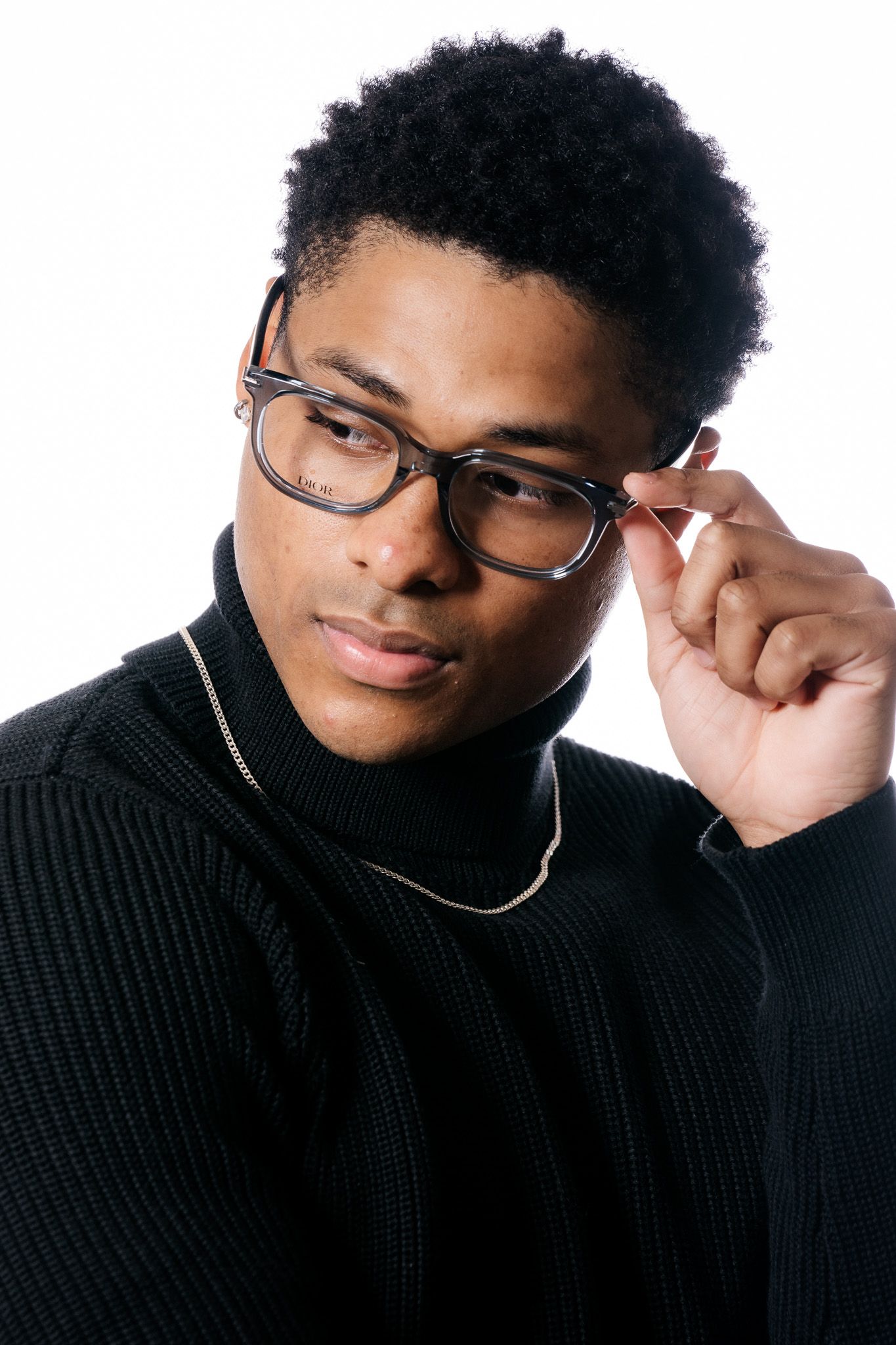 Young Black man in black turtleneck adjusting rectangular glasses with gold chain necklace on white background.